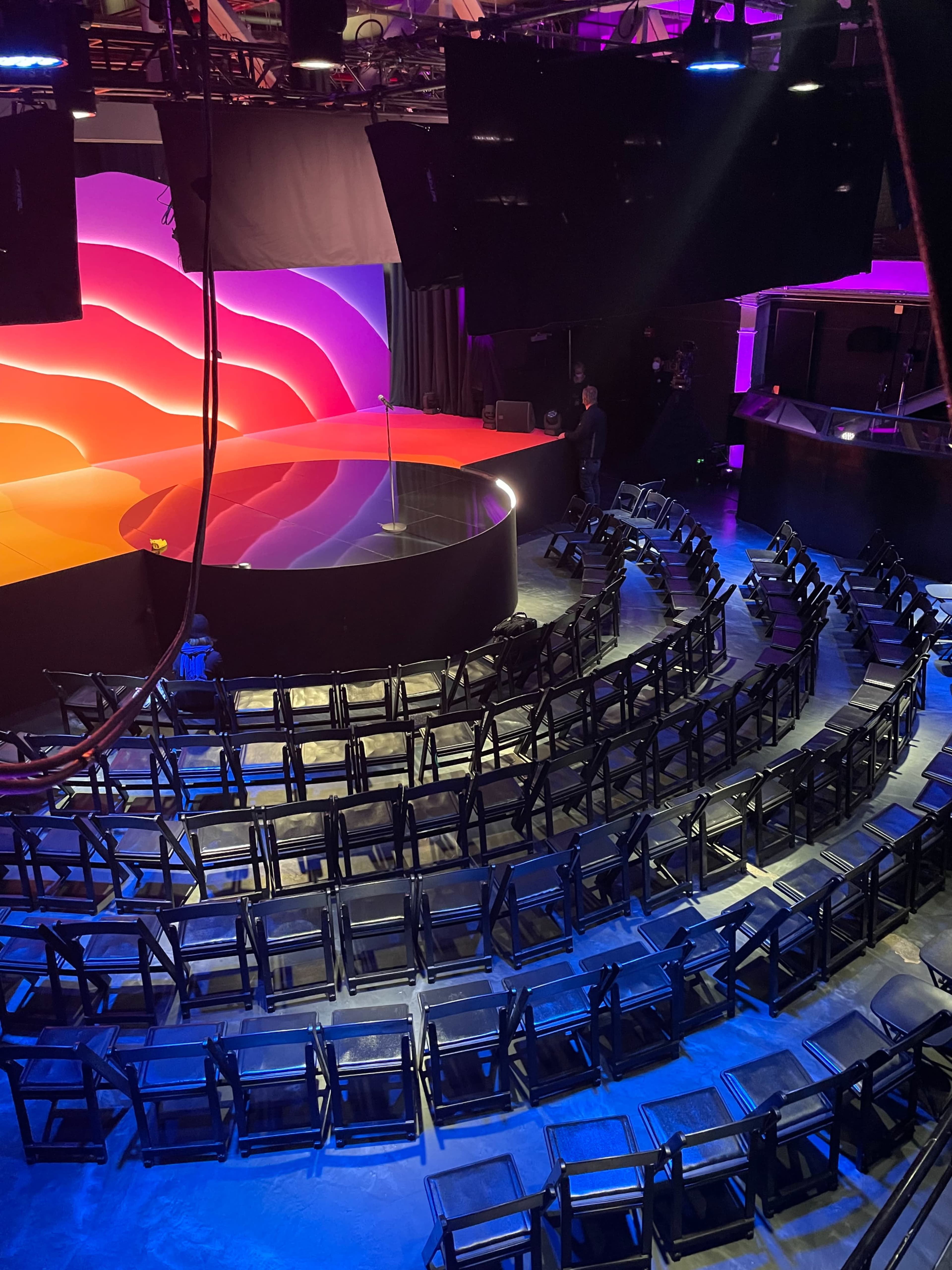 The image shows a brightly lit stage with a circular platform and rows of empty black chairs arranged in a semi-circle.