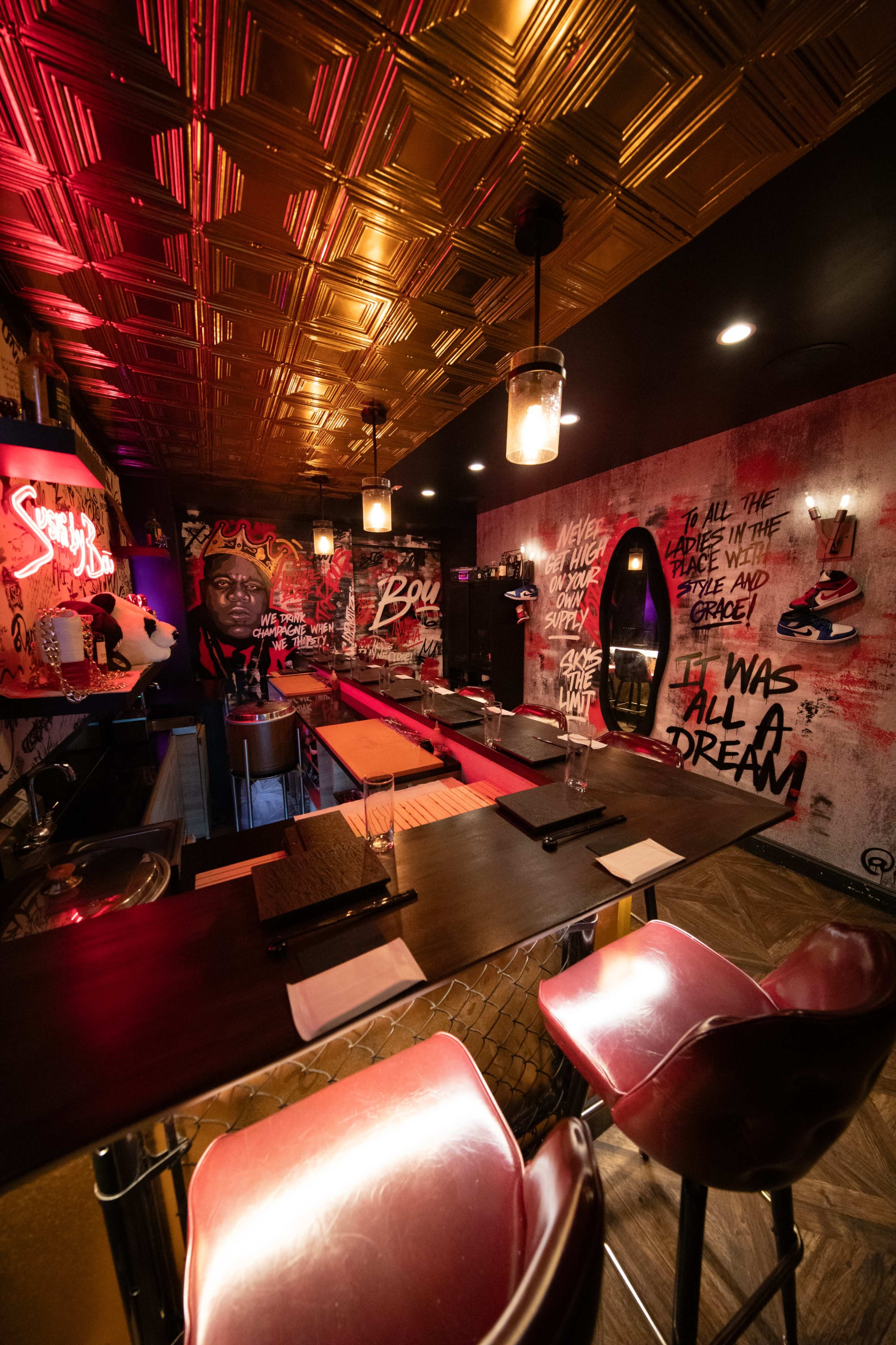 The image shows a vibrant bar interior featuring graffiti artwork, illuminated by warm lighting and furnished with red stools and wooden countertops.