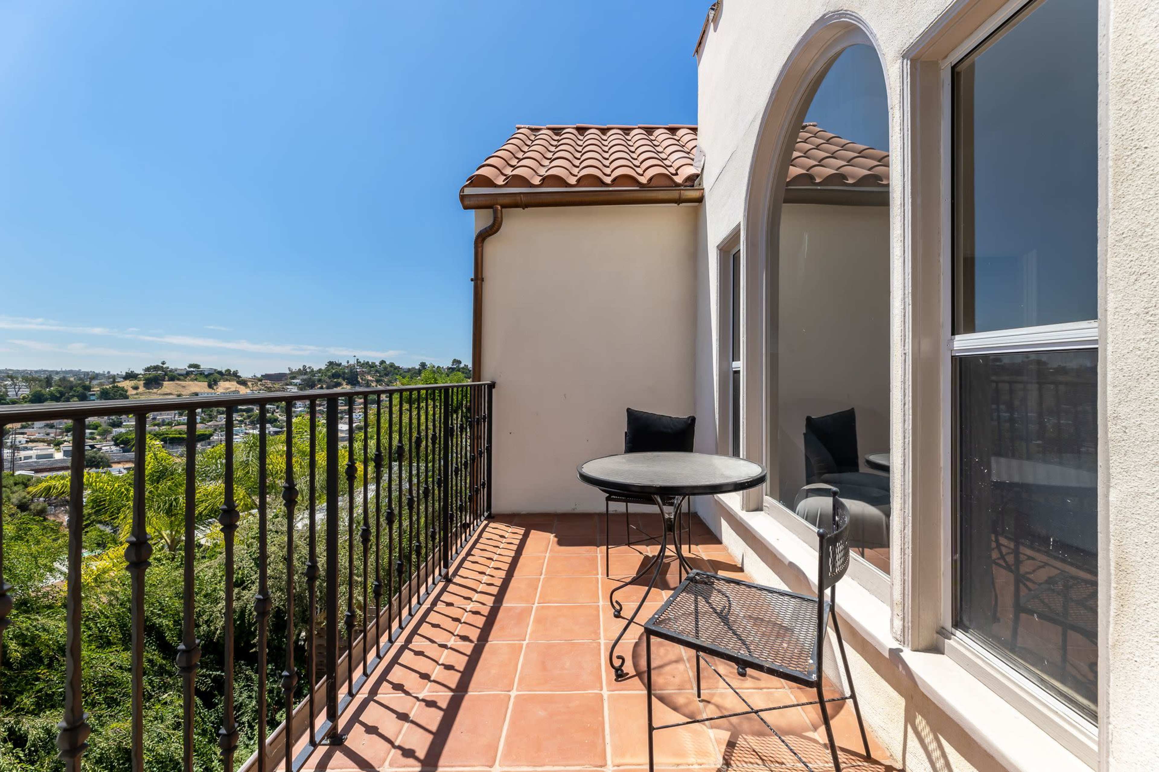 A small balcony features a round table and two chairs, overlooking a landscape with distant hills and a clear blue sky.