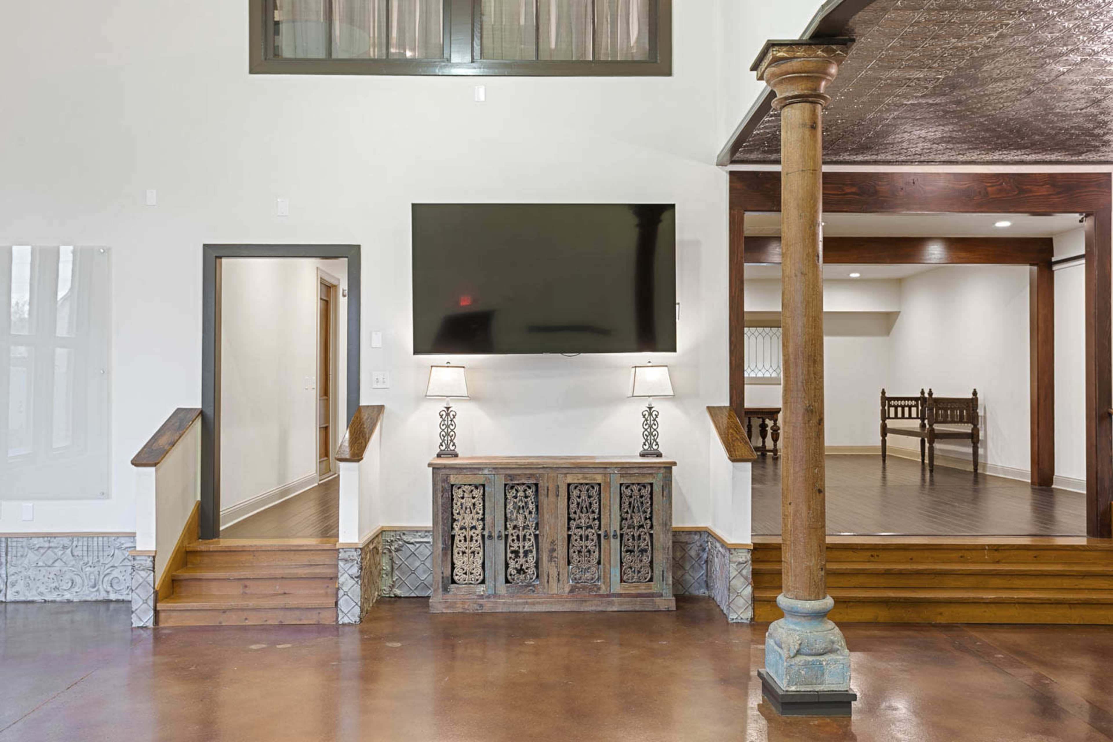 A spacious interior with a mounted television above a wooden console table, flanked by two lamps, and featuring stairs leading to a doorway on the left.