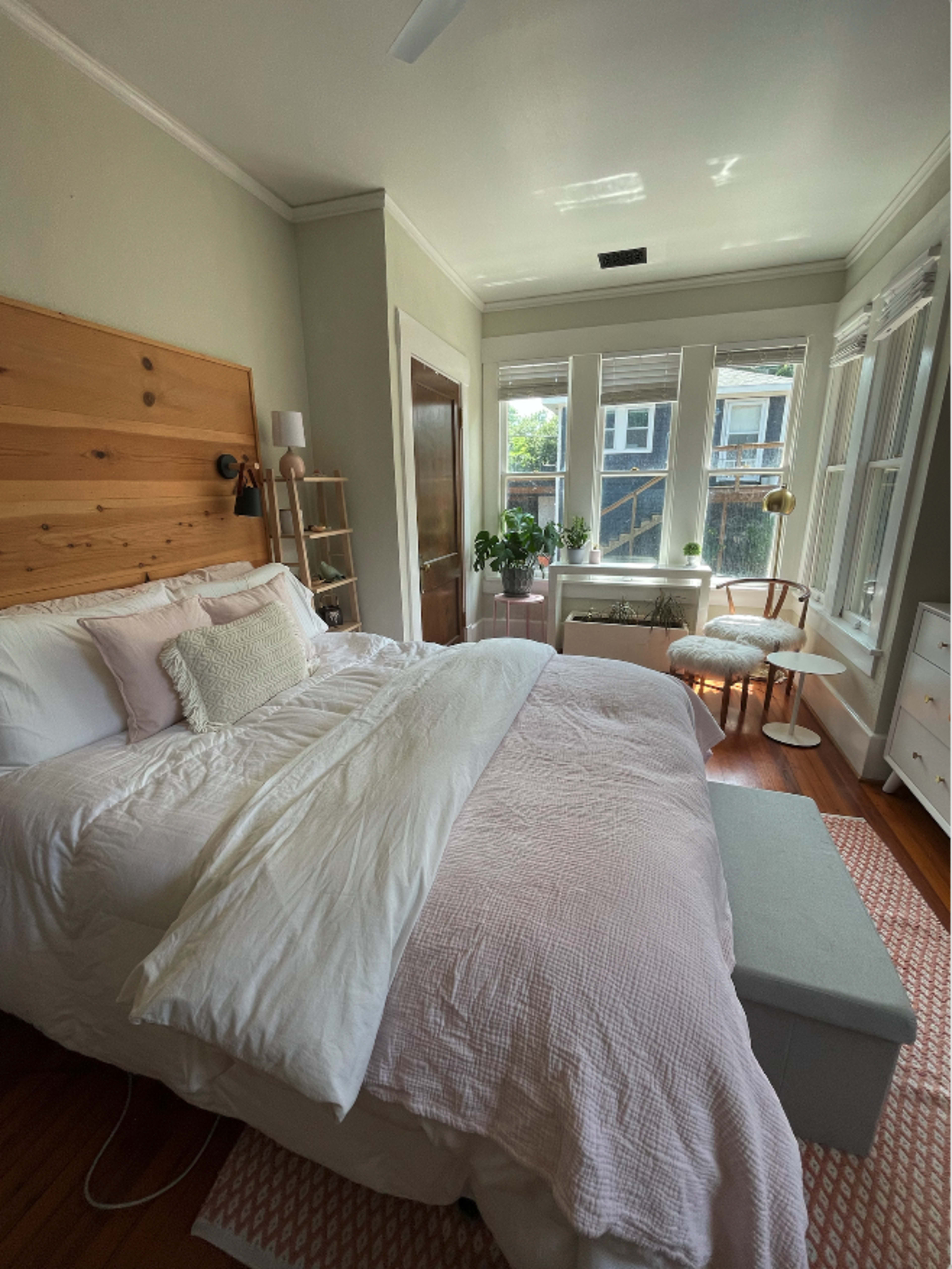 A cozy bedroom features a wood-paneled accent wall, a neatly made bed with light bedding, and large windows allowing natural light to fill the space.