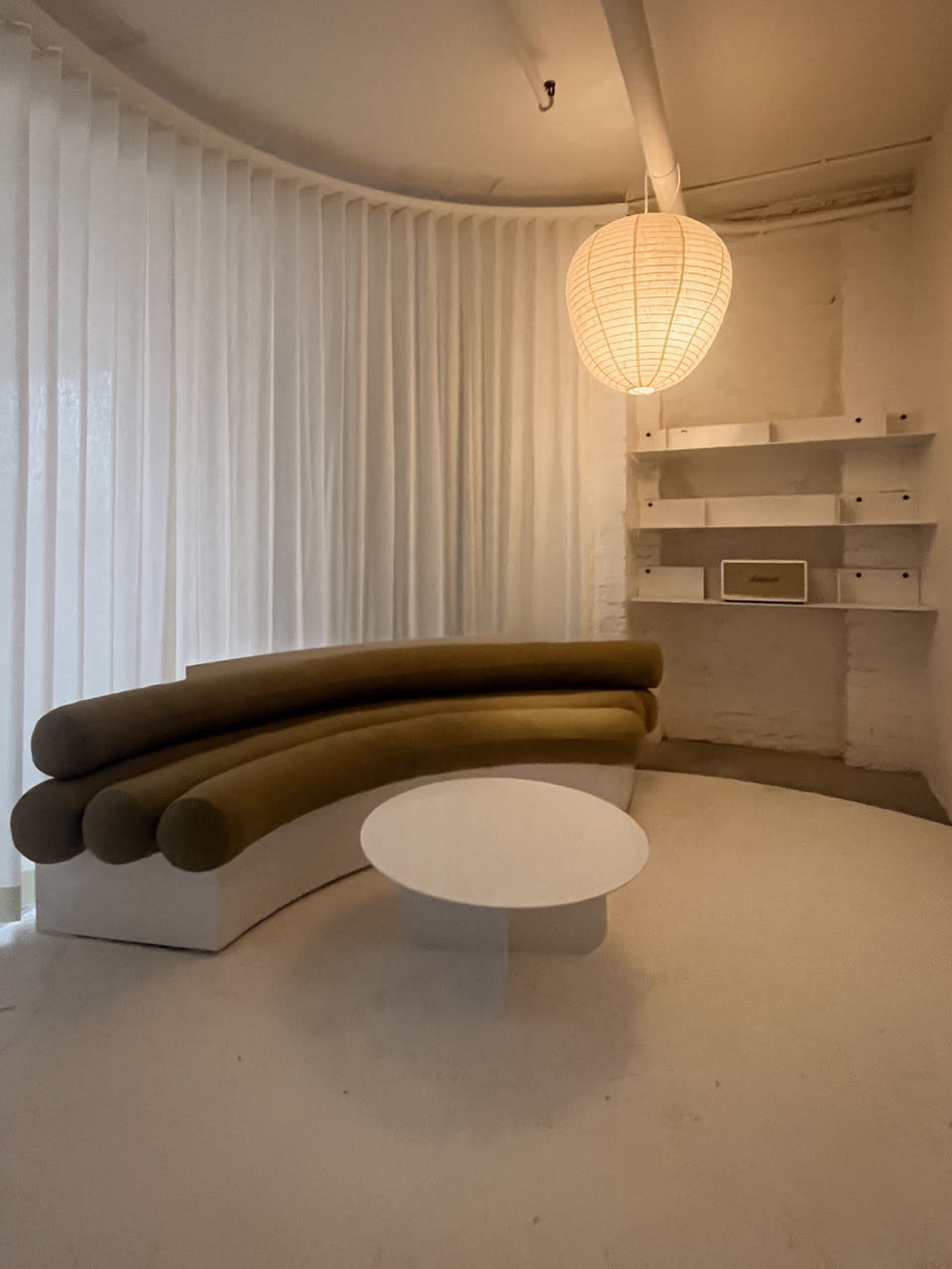 The image shows a minimalist interior featuring a curved brown sofa, a round white coffee table, and a pendant light hanging from the ceiling, with sheer curtains in the background.