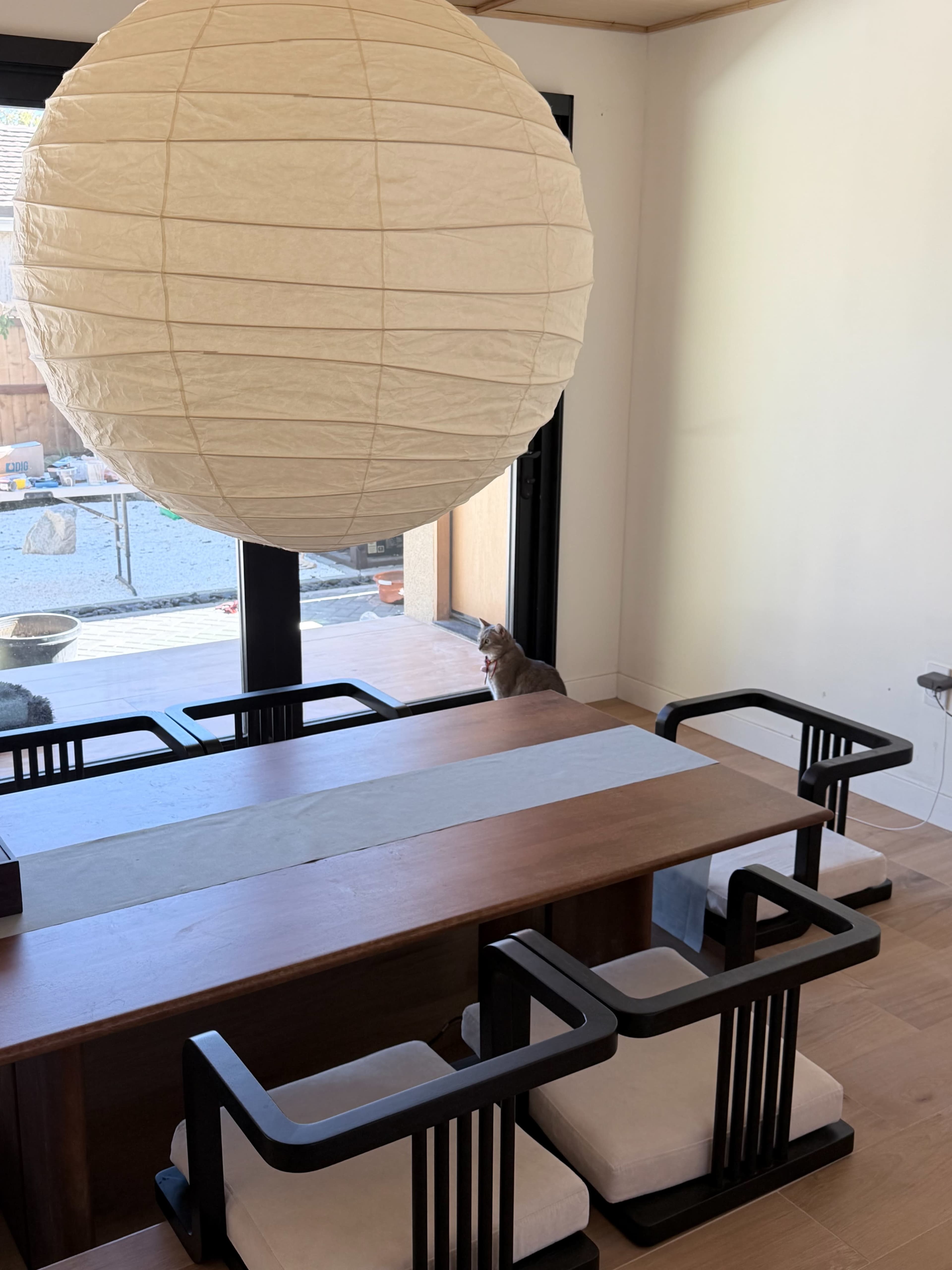 A large paper lantern hangs above a wooden dining table surrounded by black-framed chairs, with a cat sitting near a sliding glass door that opens to an outdoor area.