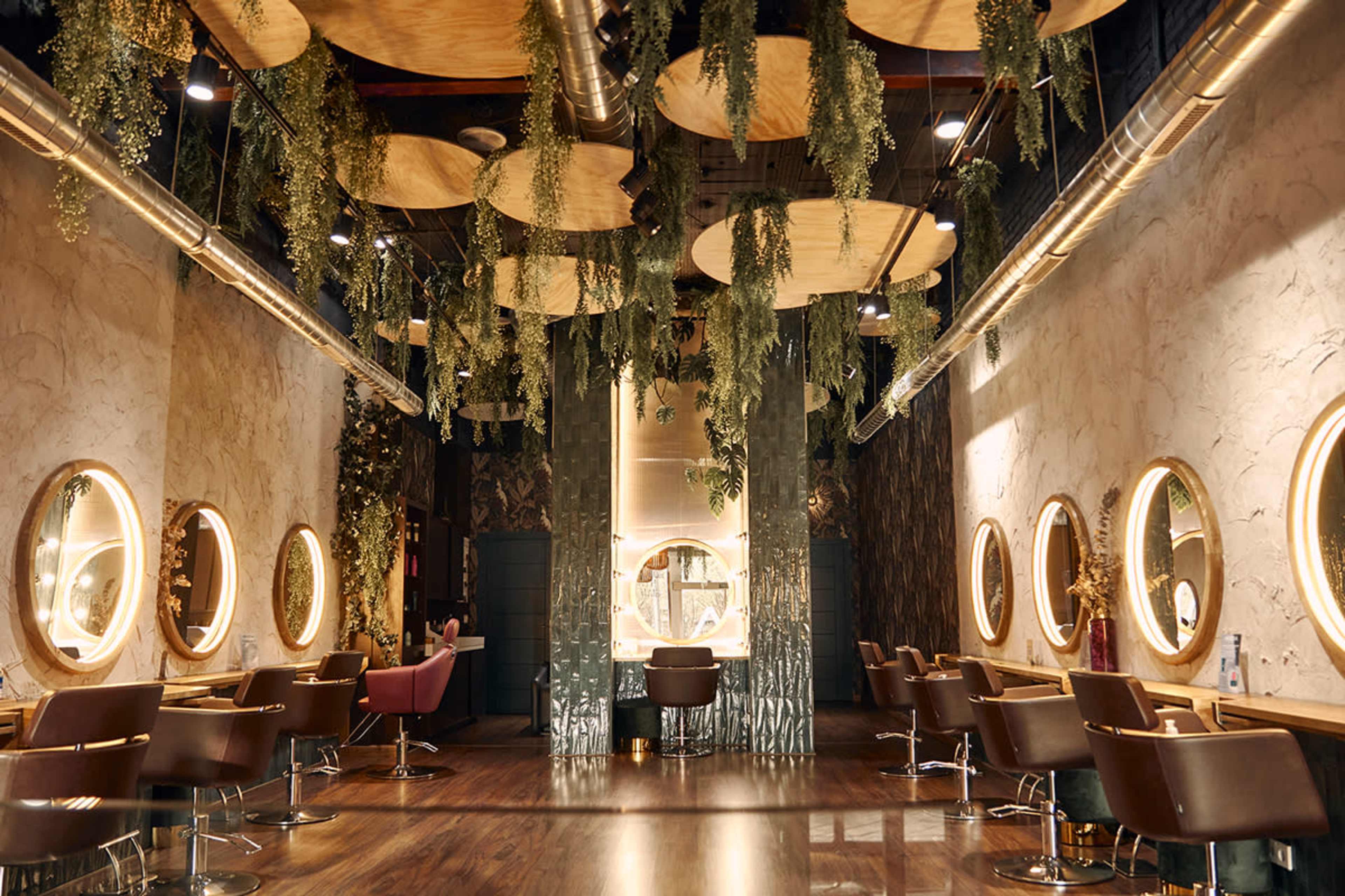 The image shows a modern salon interior with circular mirrors, wooden accents, and suspended greenery above the styling stations.