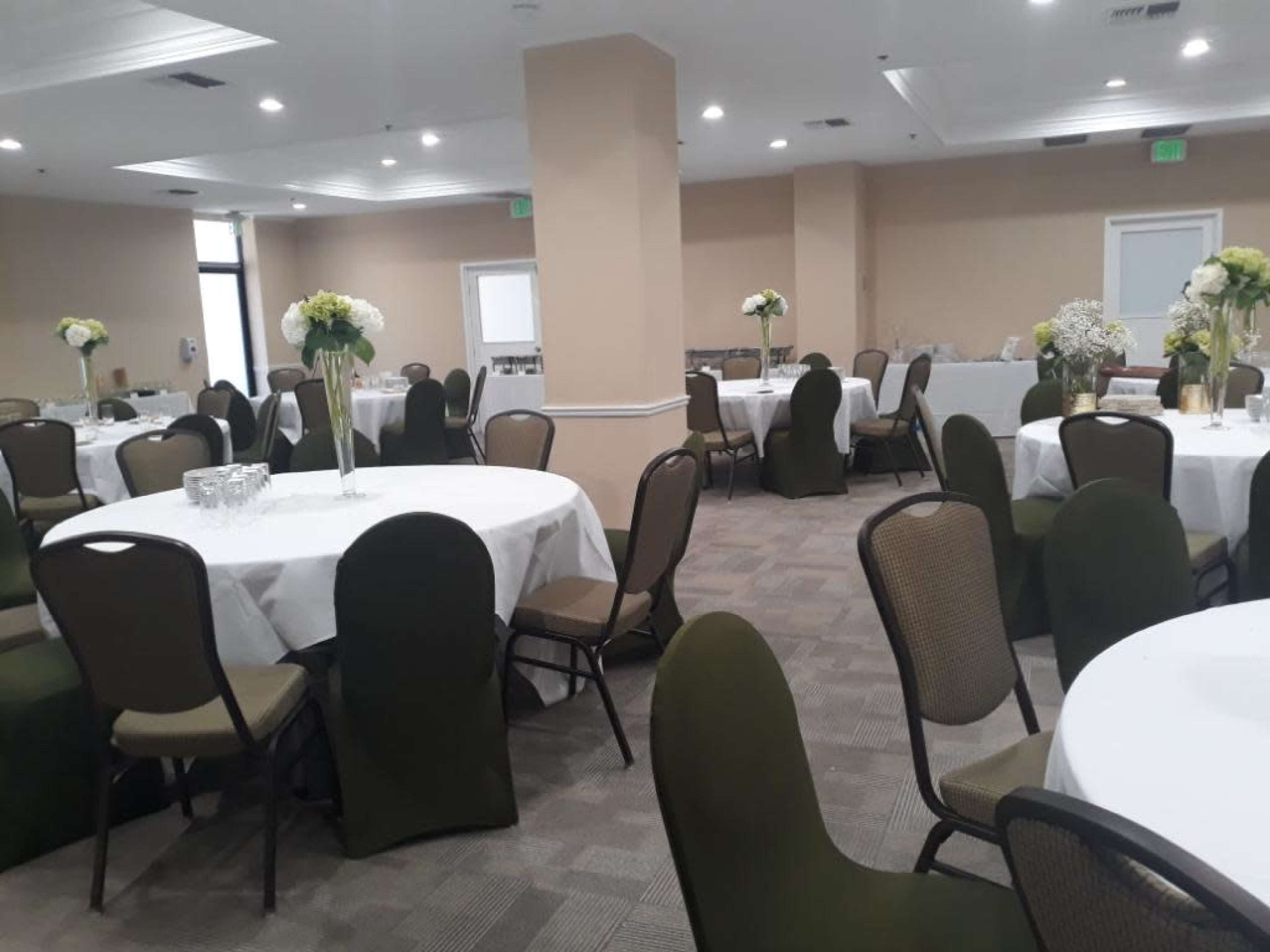 A well-lit banquet room with several round tables covered in white tablecloths, surrounded by brown chairs with green cushions and decorative floral centerpieces.