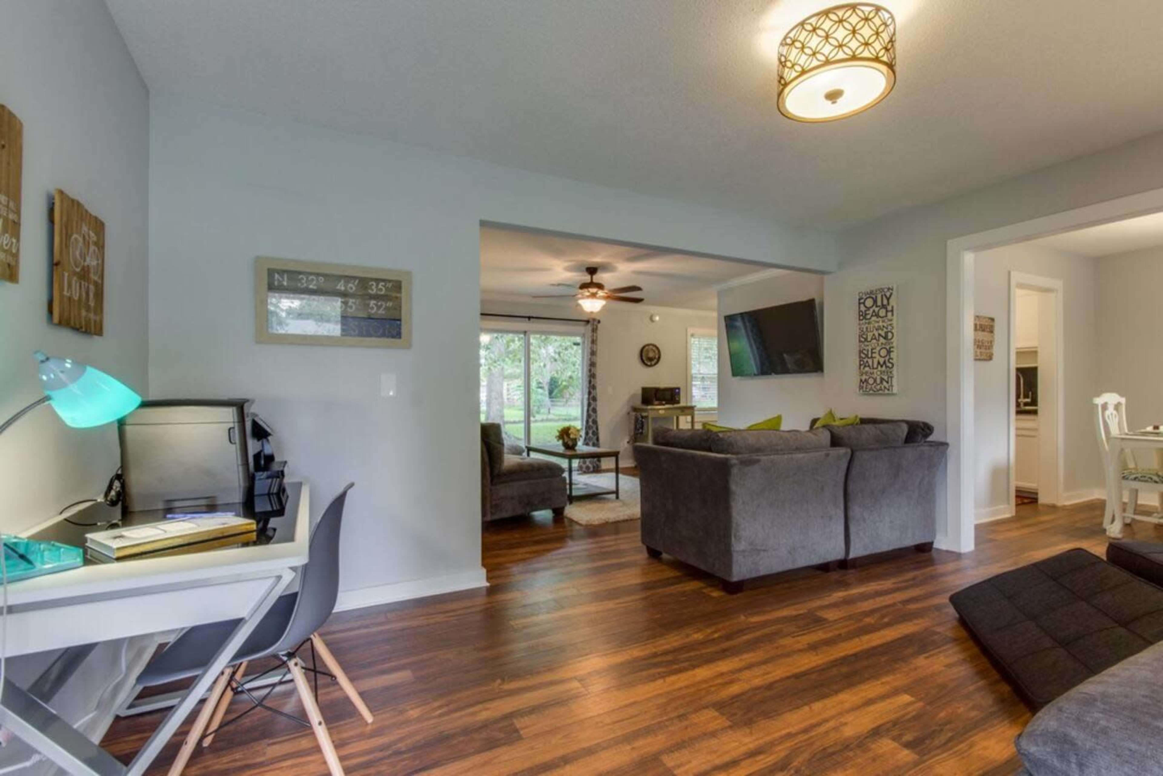 A modern living room features gray sofas, a wooden floor, and natural light coming through large windows, with a small workspace in the foreground.