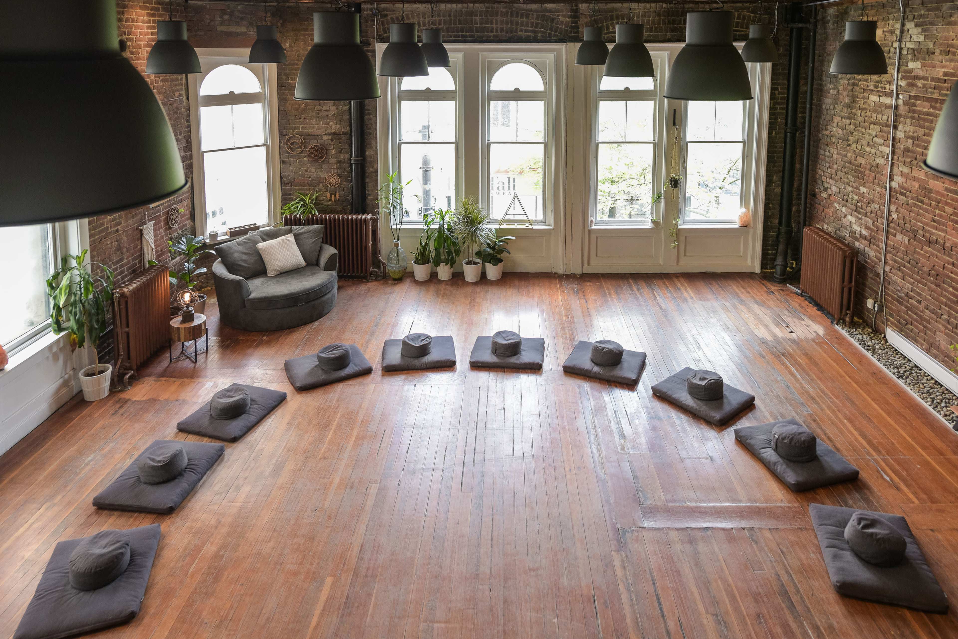 An empty meditation studio with a circular arrangement of gray cushions on a wooden floor, a cozy armchair in the corner, and large windows letting in natural light.