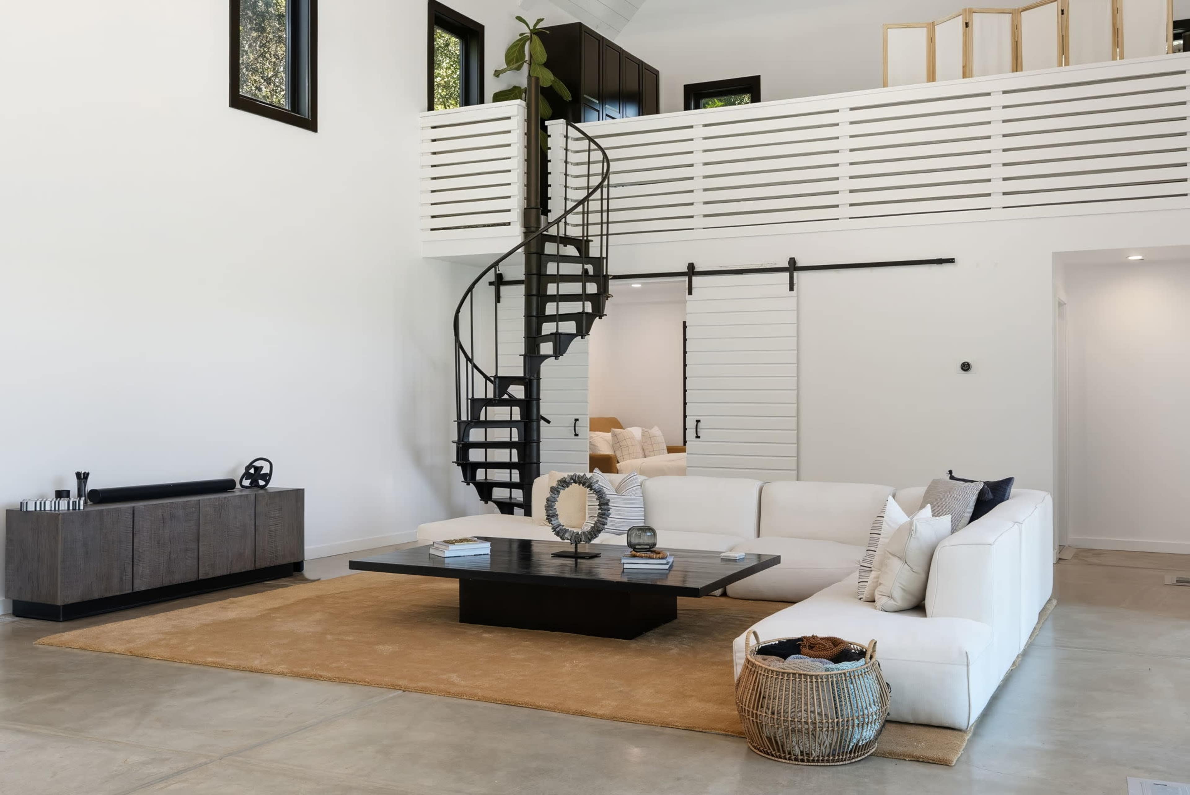 The image features a modern living room with a white sectional sofa, a large circular coffee table, a spiral staircase, and minimalist decor on a concrete floor.