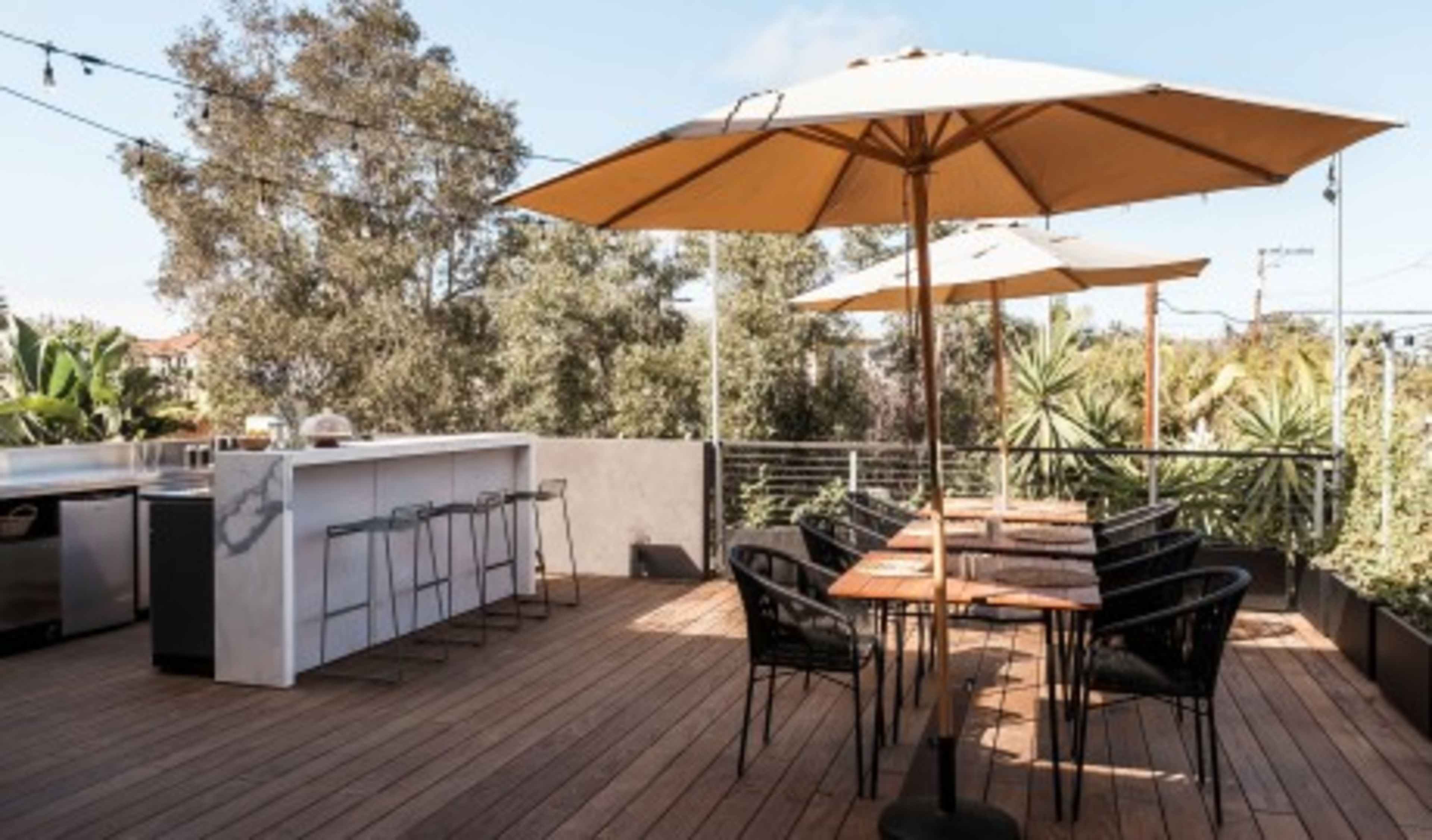 A rooftop terrace features wooden decking with a bar, high stools, dining tables, and large umbrellas surrounded by greenery.