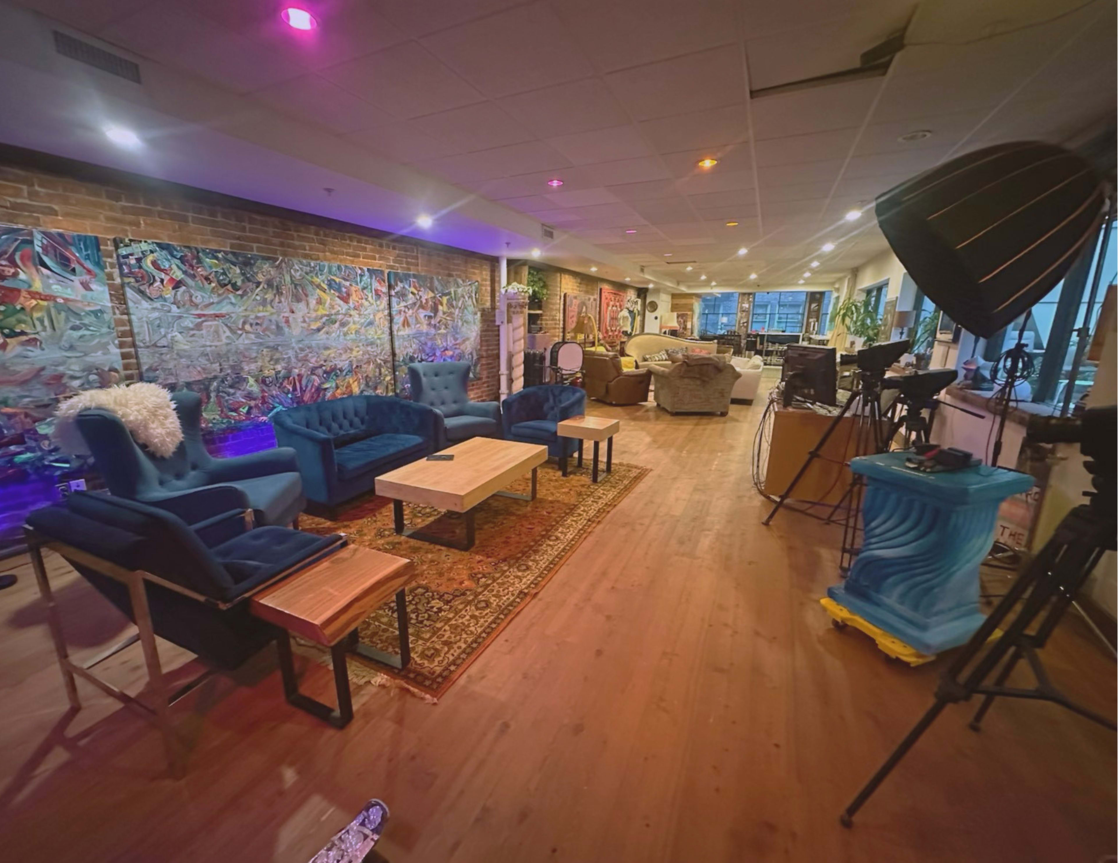 The image shows a spacious, modern lounge area with blue armchairs, a wooden table, artistic wall decor, and various seating arrangements in a well-lit environment.