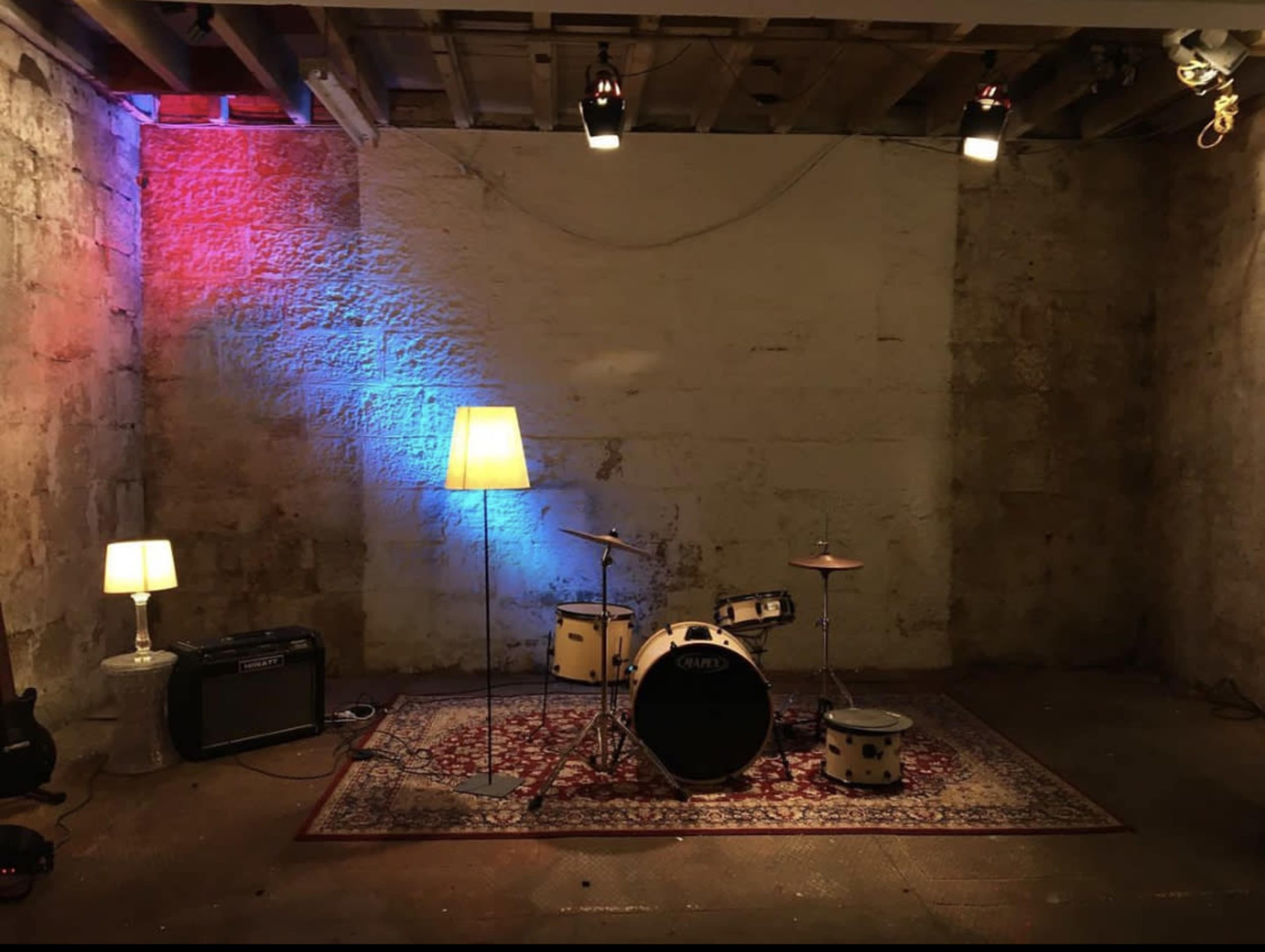 A small, dimly lit room features a drum set, two floor lamps, and an amplifier against a stone wall.