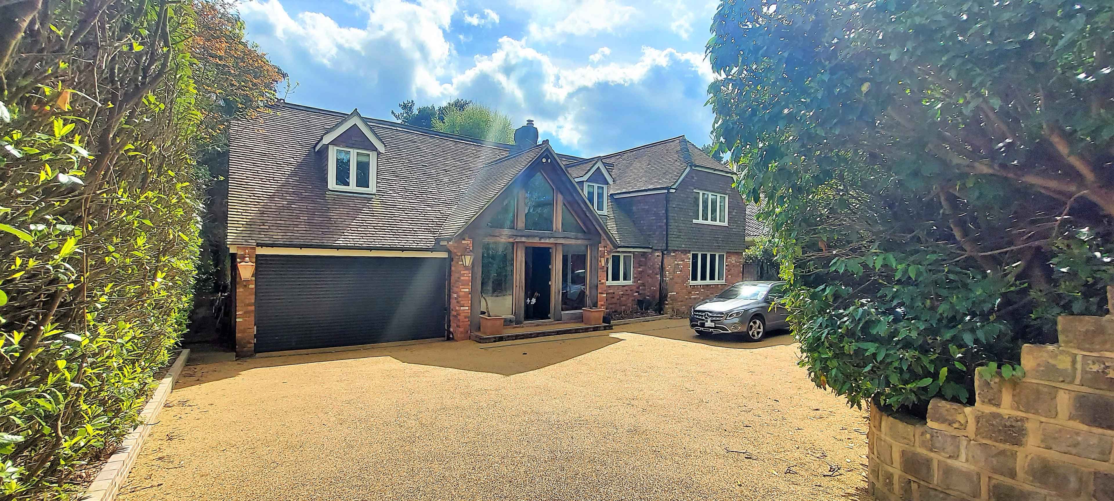 A two-story house with a large front entrance and a gravel driveway is surrounded by greenery.
