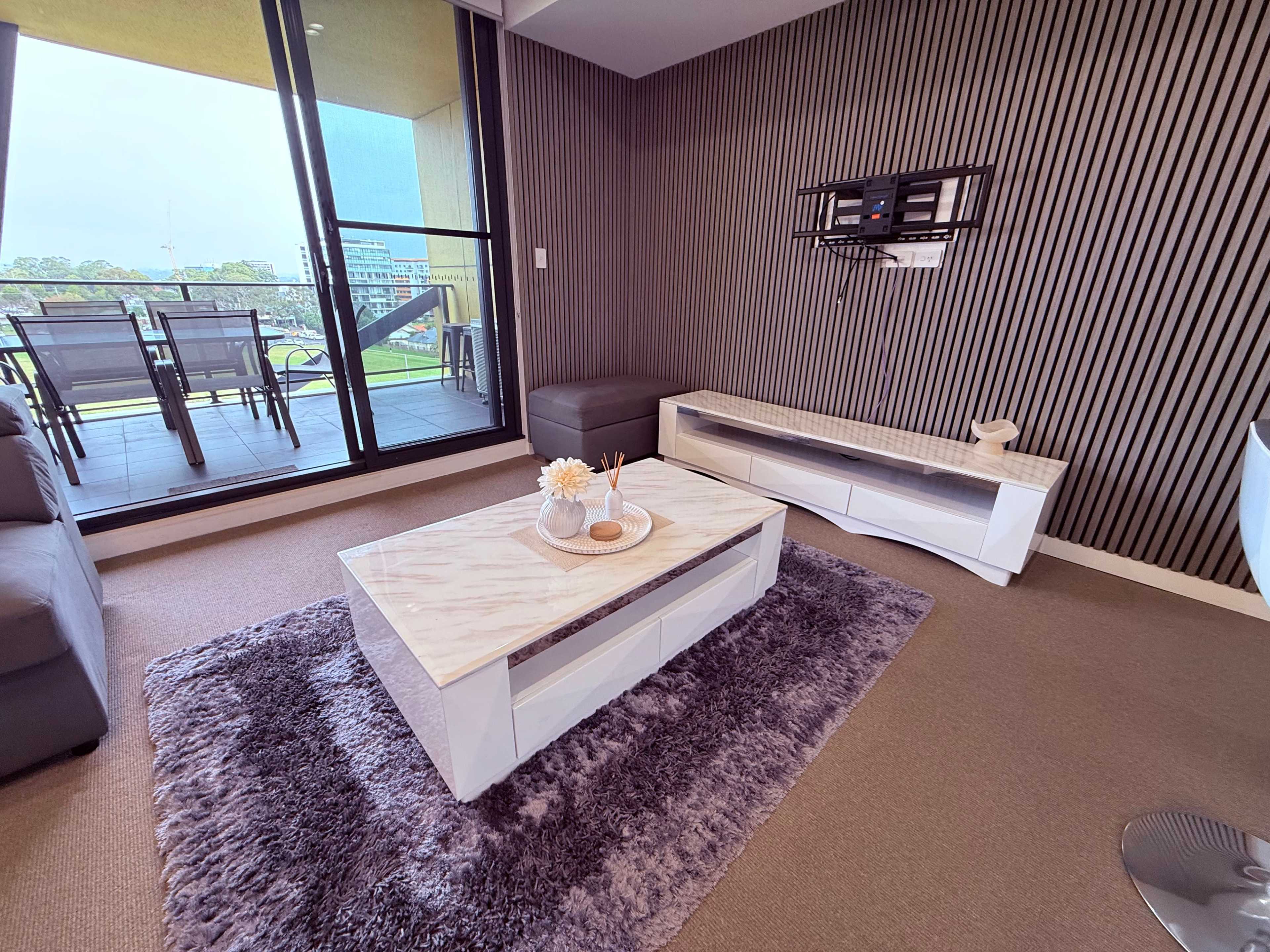 The image shows a modern living room with a marble-top coffee table, a plush rug, and large windows leading to an outdoor balcony.