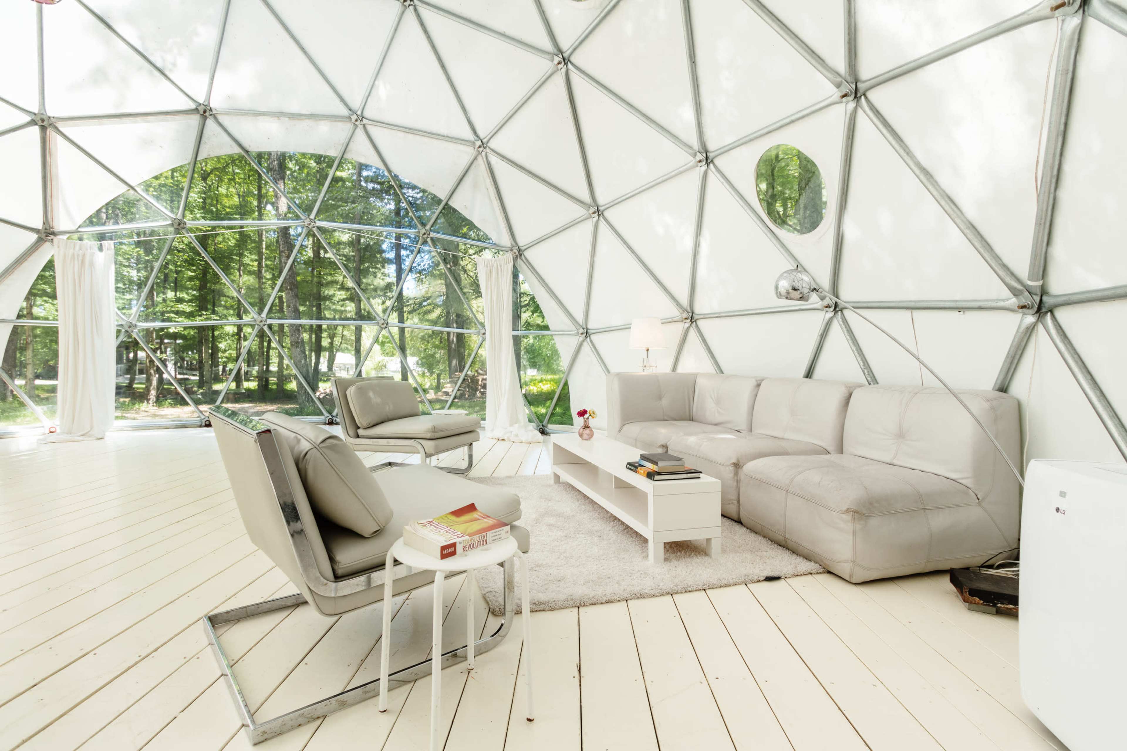 The interior of a geodesic dome features a light-colored sectional sofa, armchairs, a coffee table, and large windows overlooking a forested area.
