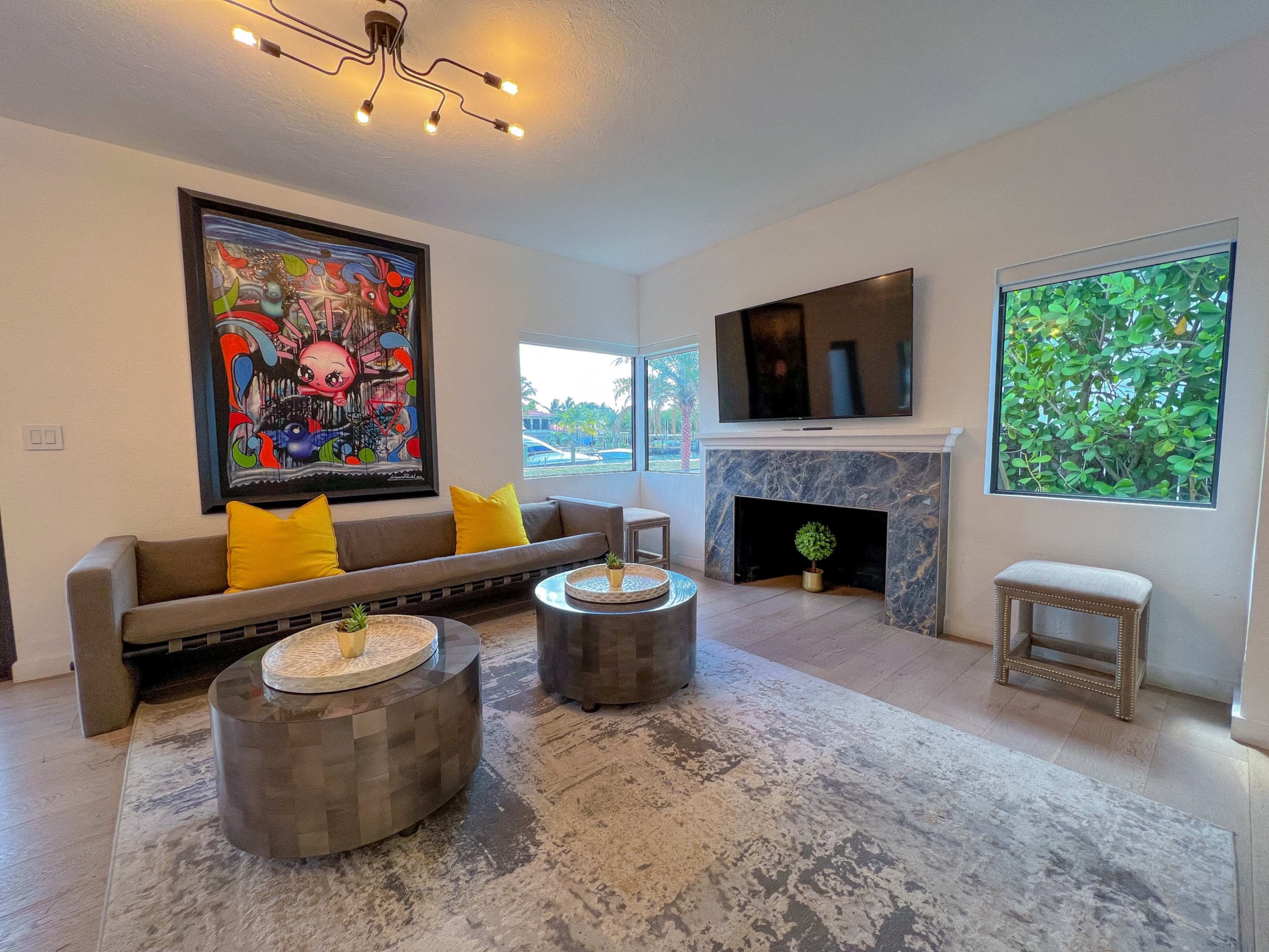 The living room features a gray sofa with yellow cushions, two circular tables, a large artwork on the wall, and a TV above a marble fireplace.