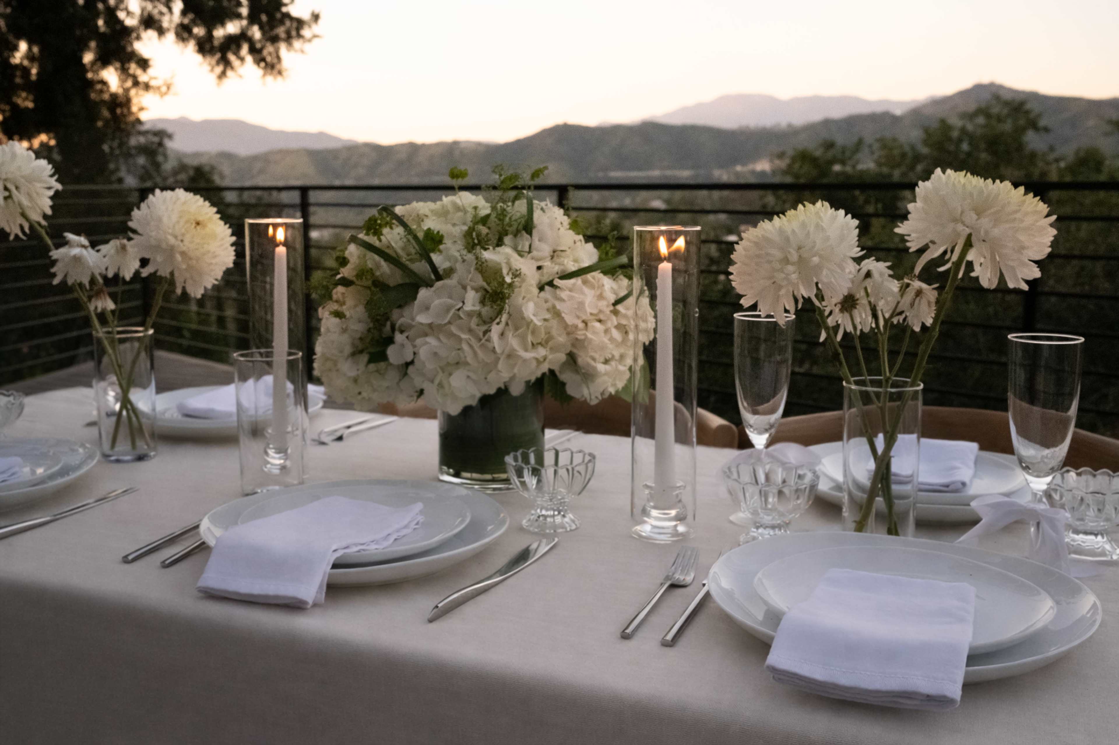 A dining table is set with plates, glasses, and a floral centerpiece, overlooking a mountainous landscape at sunset.