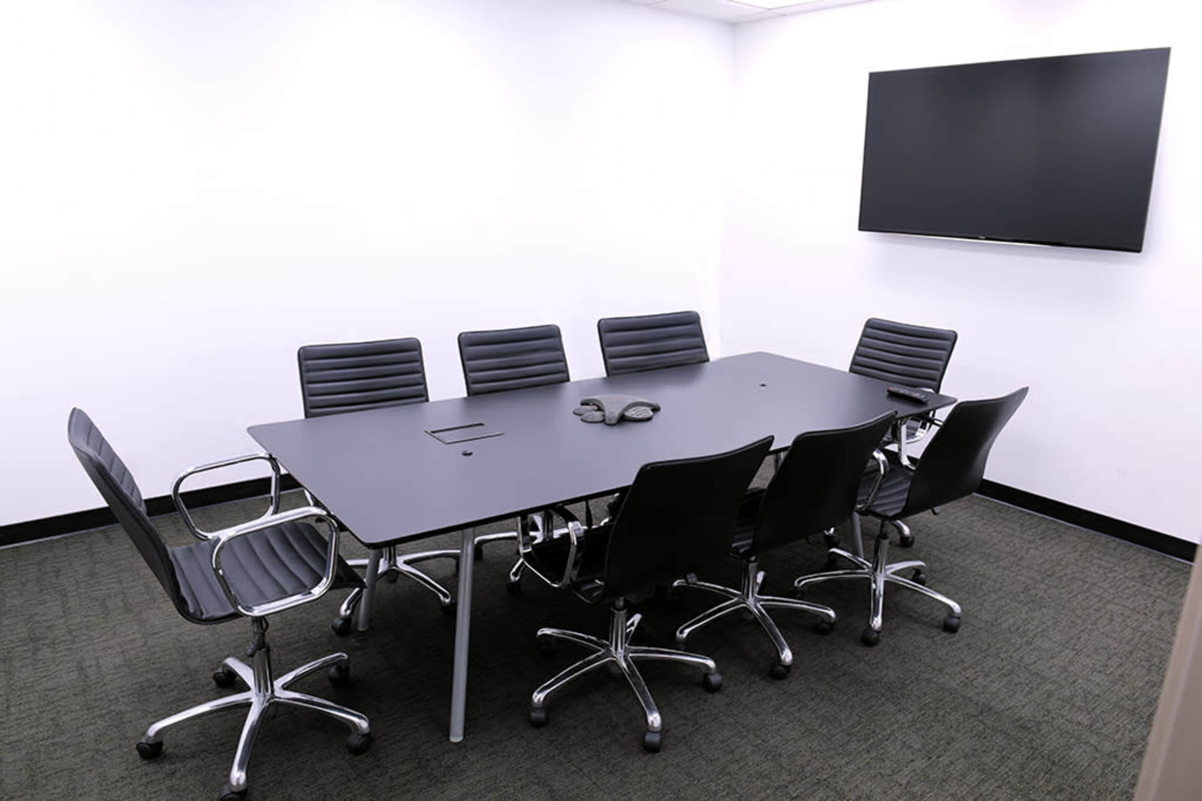 A modern conference room features a long black table surrounded by eight black swivel chairs, with a wall-mounted display screen in the background.