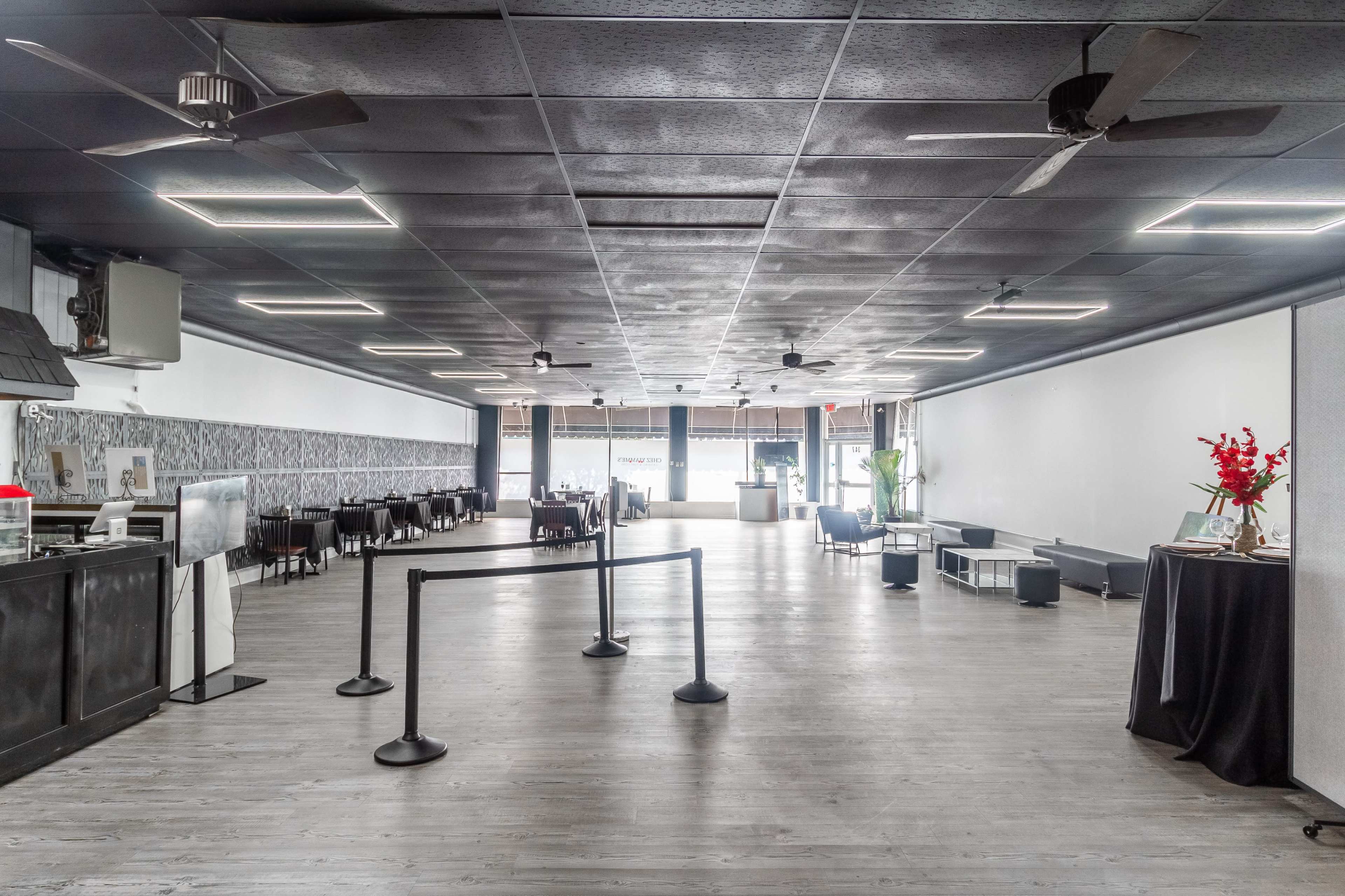 A spacious, empty commercial interior with a smooth wooden floor, ceiling fans, and minimal furnishings set against white walls and large windows.