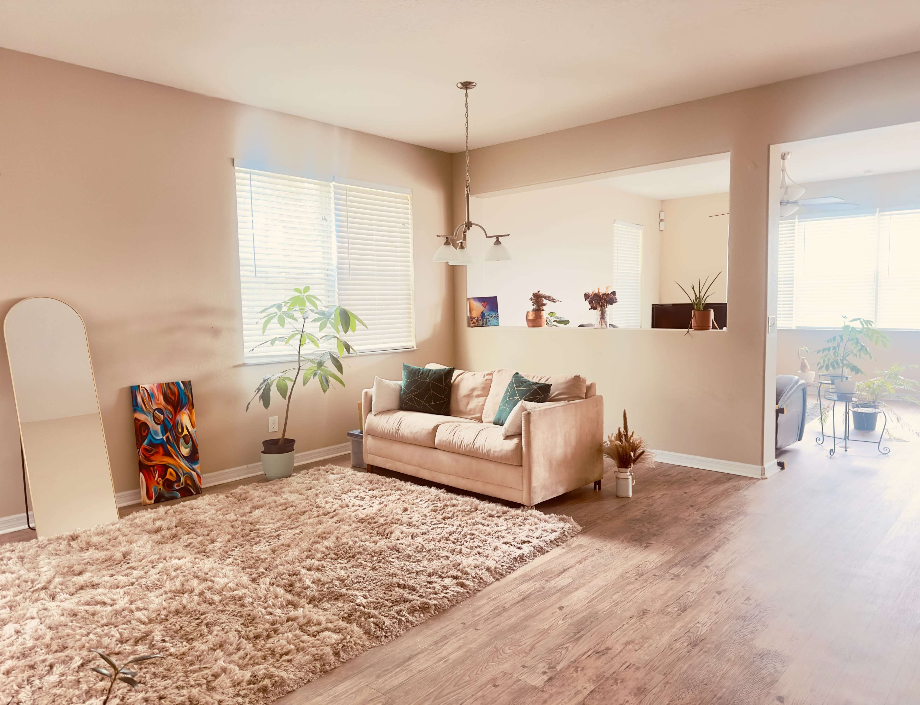 The image shows a light-filled living room with a beige sofa, a large area rug, and decorative plants, alongside a mirror and artwork on the walls.