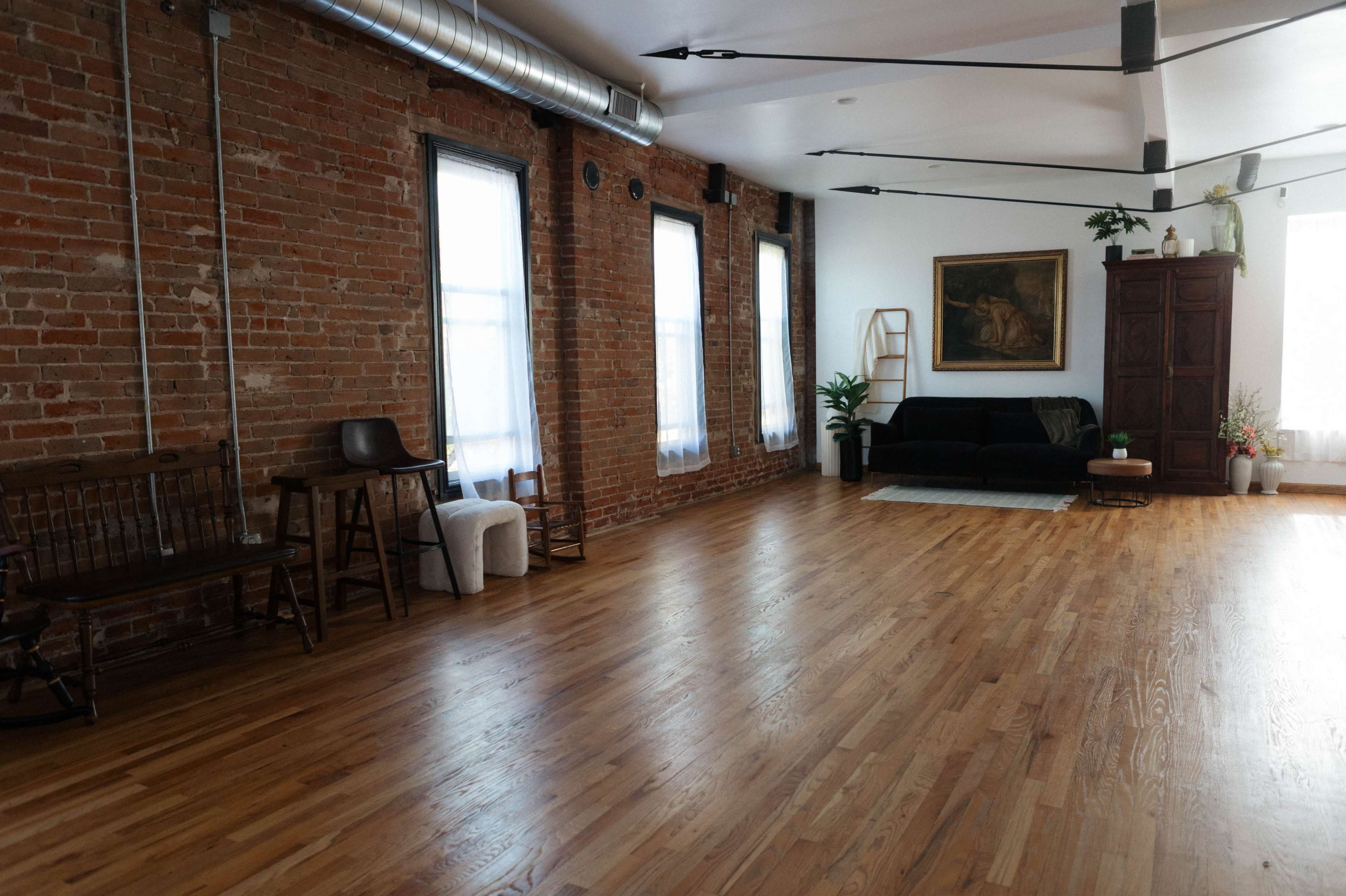 The image shows an interior space with exposed brick walls, hardwood flooring, and a black sofa, along with plants and artwork.