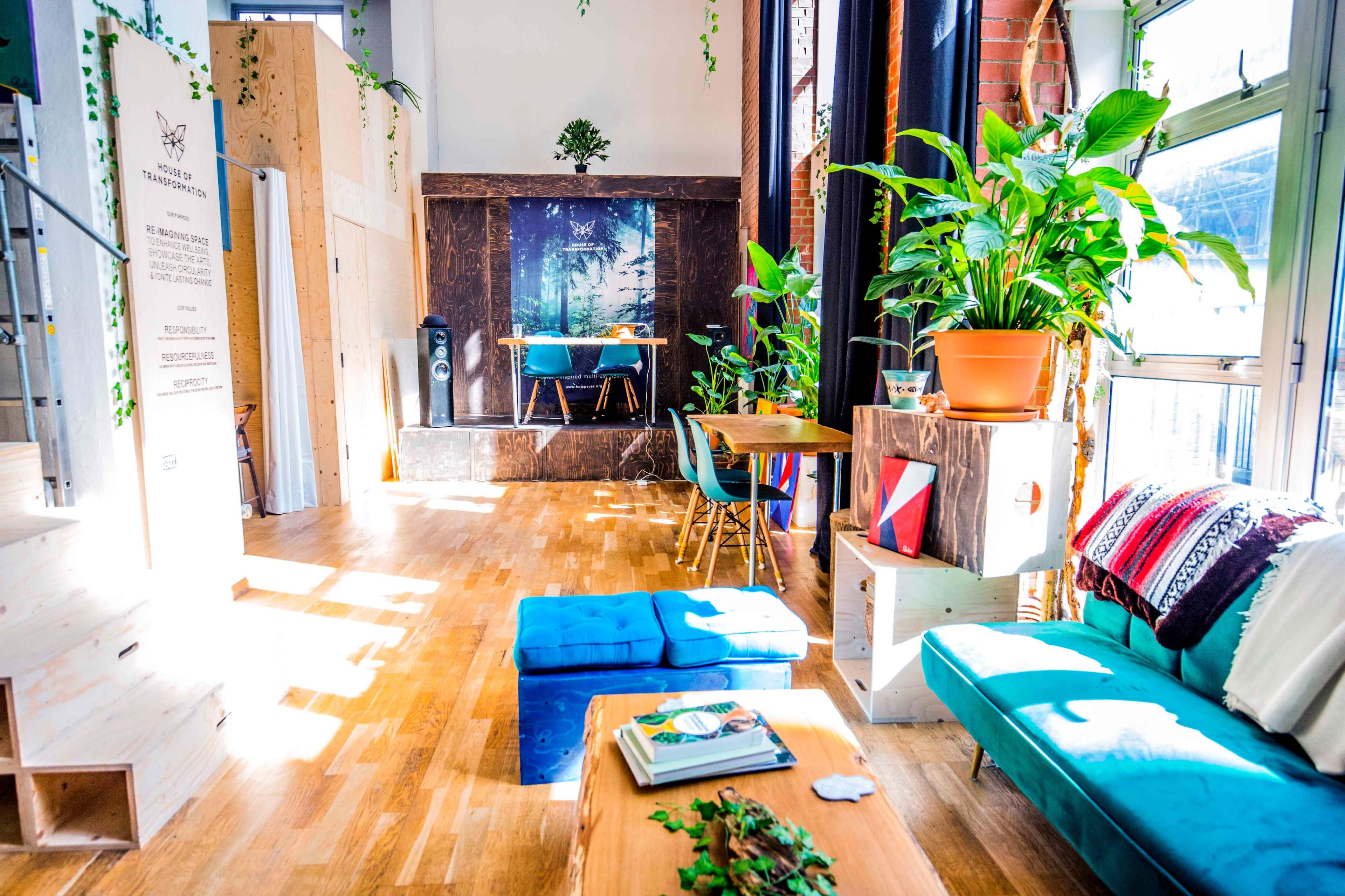 The image shows a bright and modern interior space with wooden flooring, plants, a blue couch, a wooden table, and a backdrop featuring a forest scene.