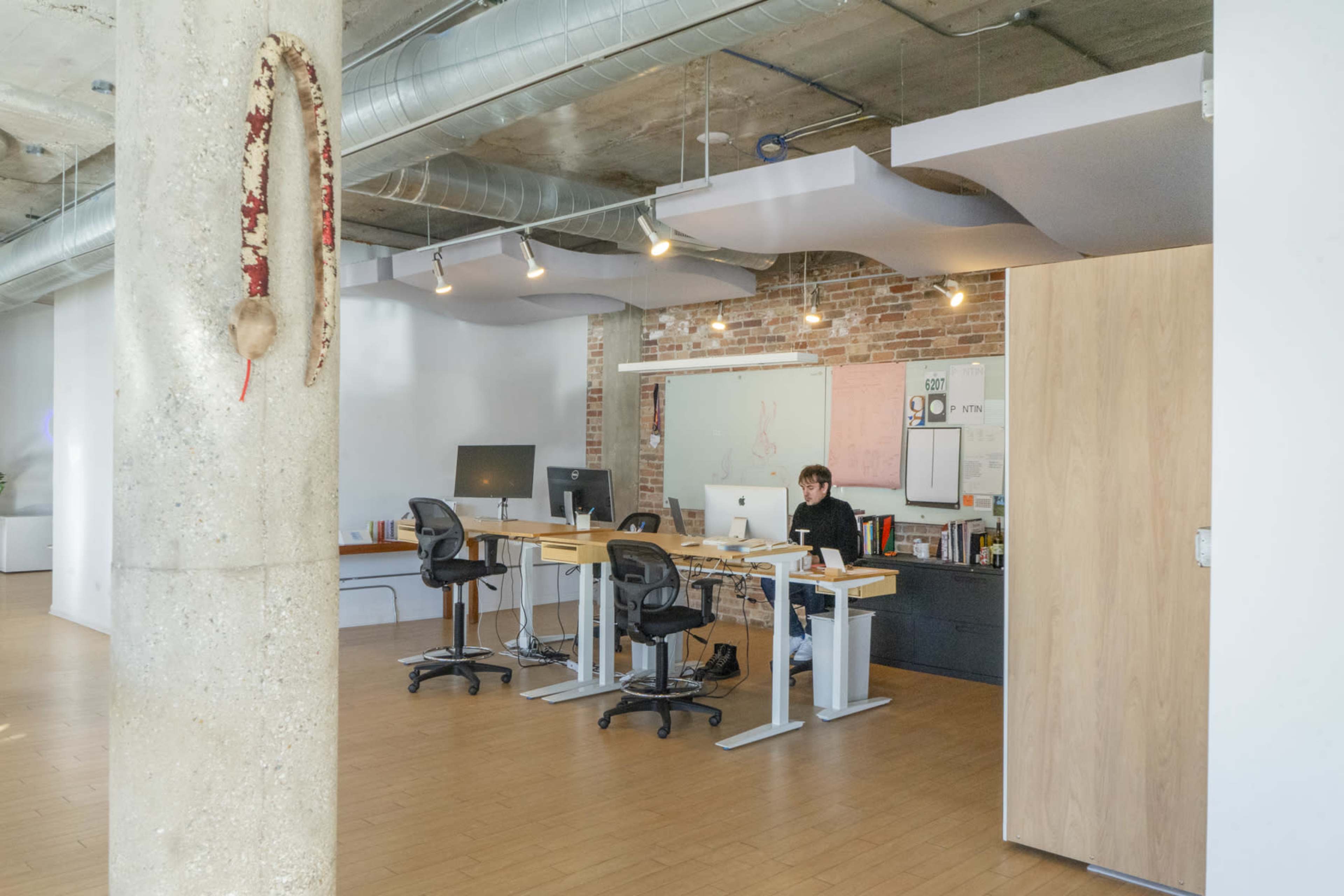 An office space features a worker at a desk with computers, surrounded by wooden flooring, exposed brick walls, and modern lighting.