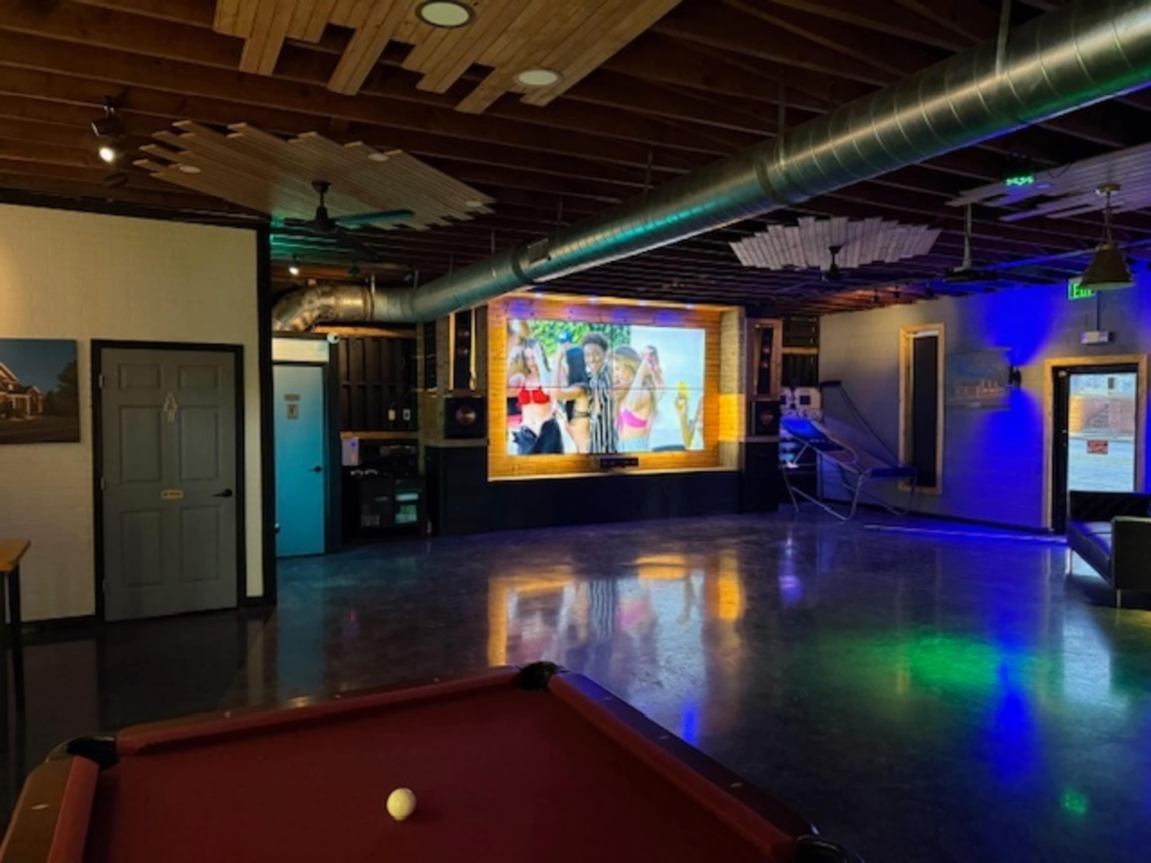 The image shows a spacious room with a pool table in the foreground and a large screen displaying colorful images on the wall.