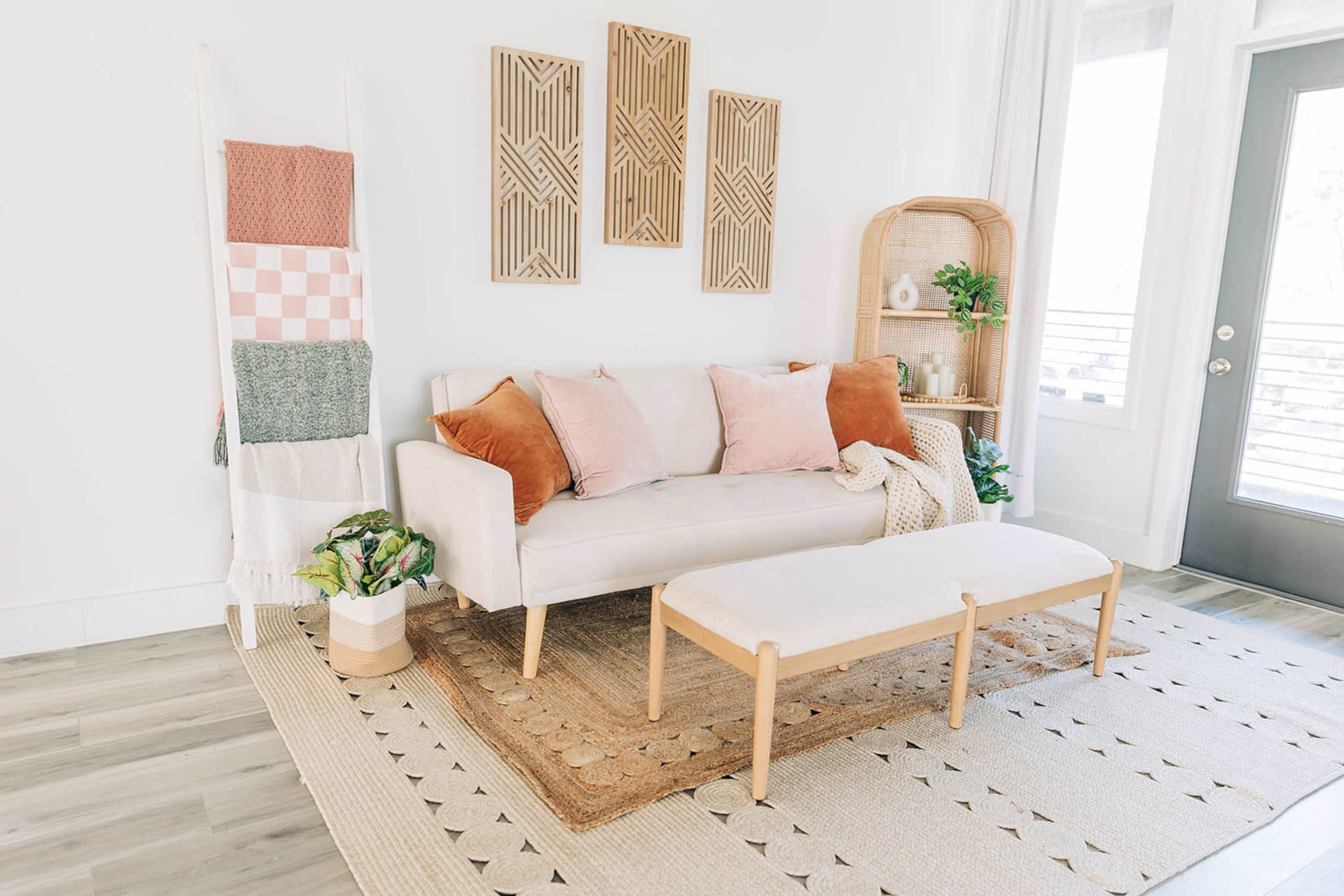 A light-colored living room features a sofa with decorative pillows, a bench, wall art, and a woven basket holding plants.