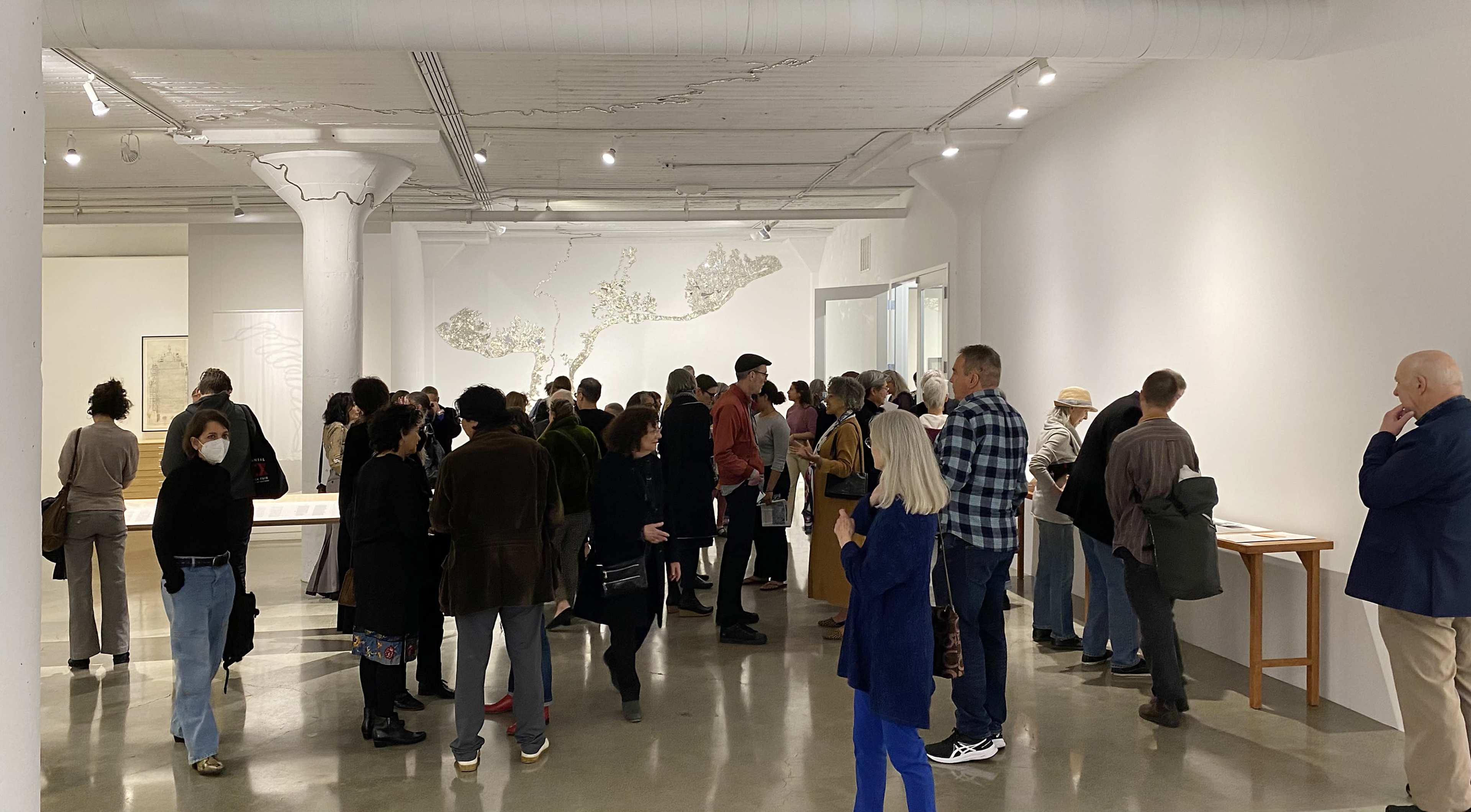 A large group of people is gathered in an art gallery, looking at exhibits and conversing with one another.
