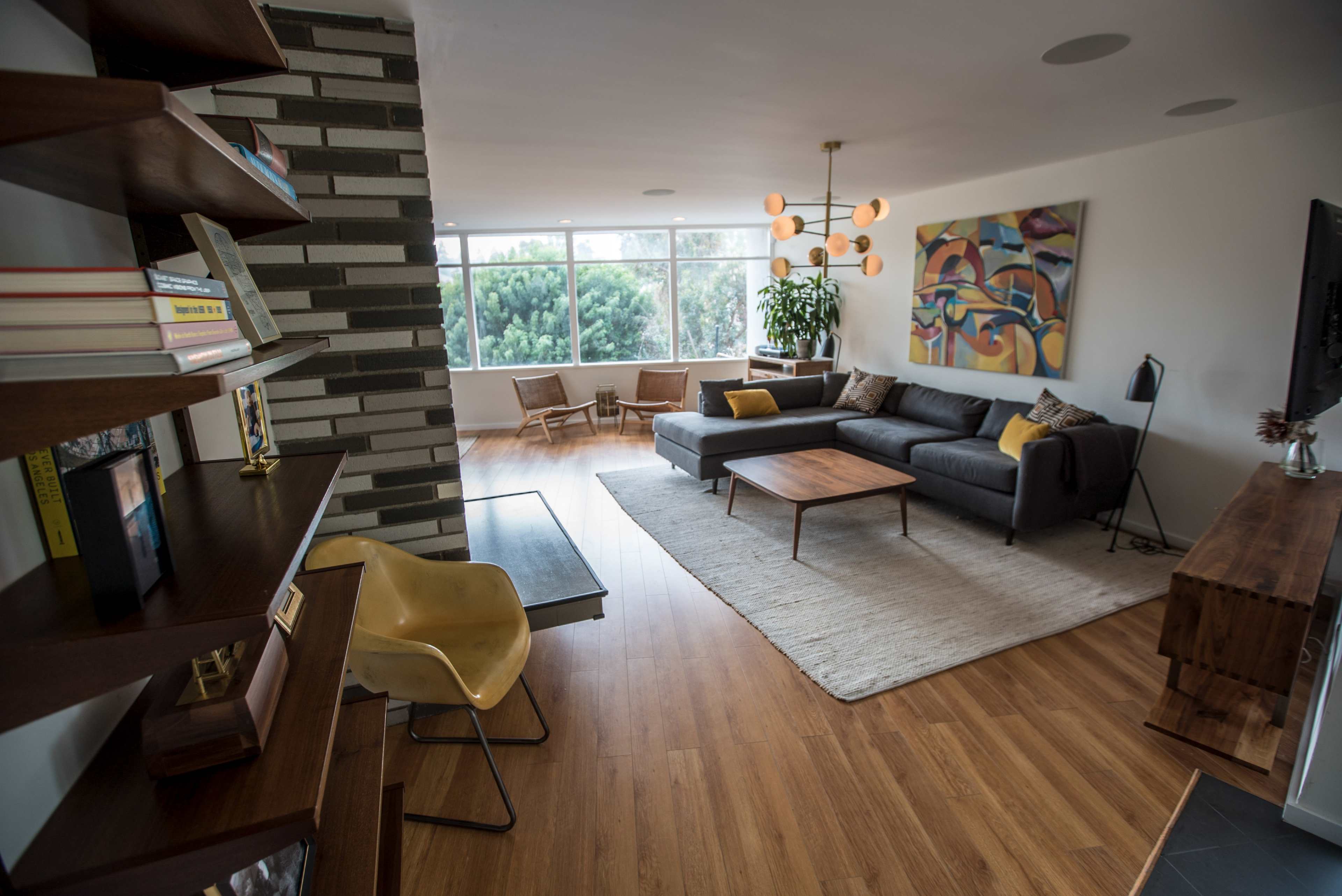 The image shows a modern living room featuring a gray couch, a wooden coffee table, a bookshelf, and large windows that allow natural light to enter.