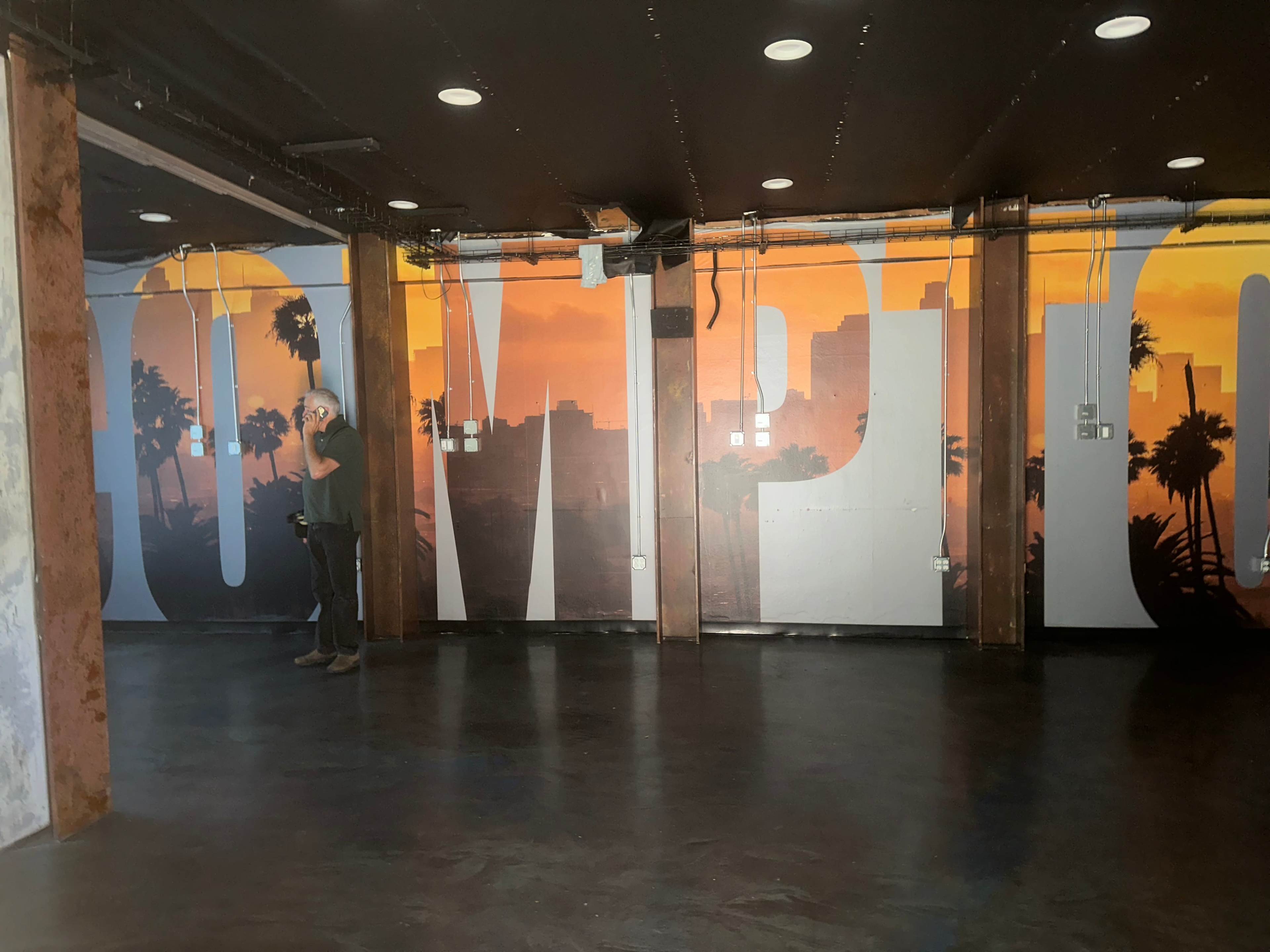 A person stands in a room with large "COMPTON" lettering on the wall, illuminated by a sunset backdrop featuring palm trees.