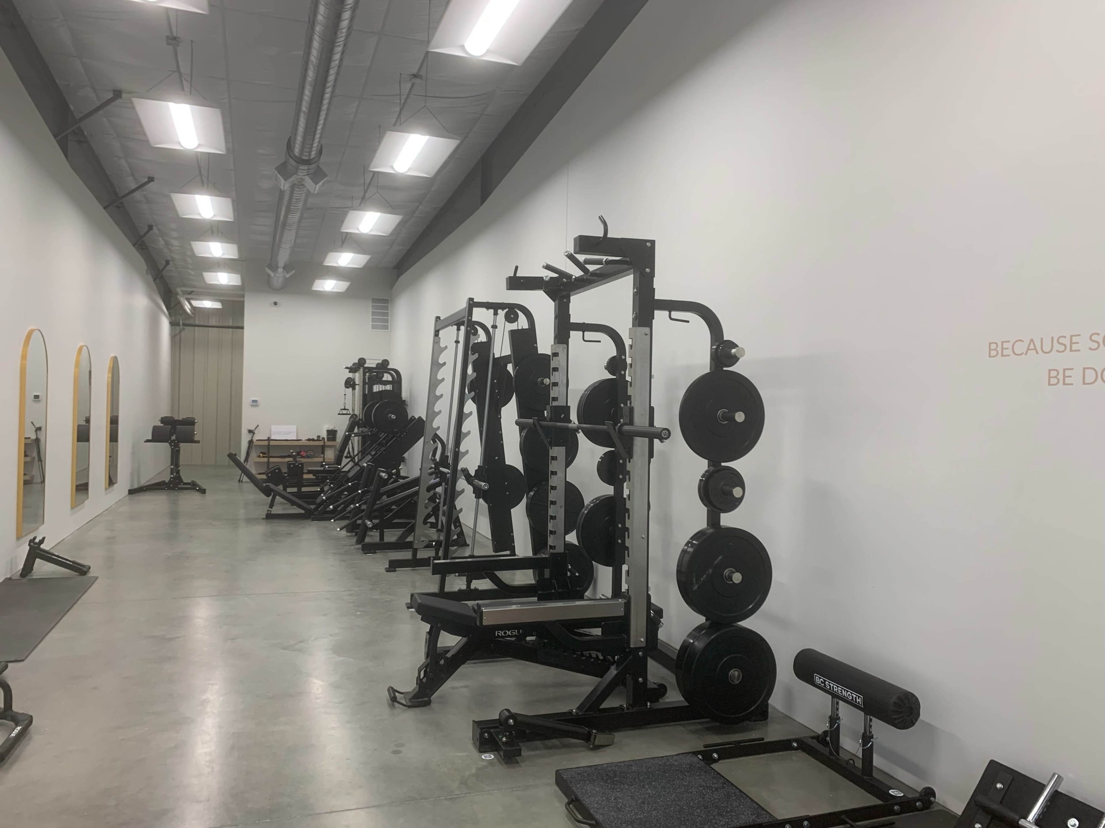 The image shows a fitness facility featuring weightlifting equipment, including benches and squat racks, along a well-lit, minimalist space with mirrors on one side.