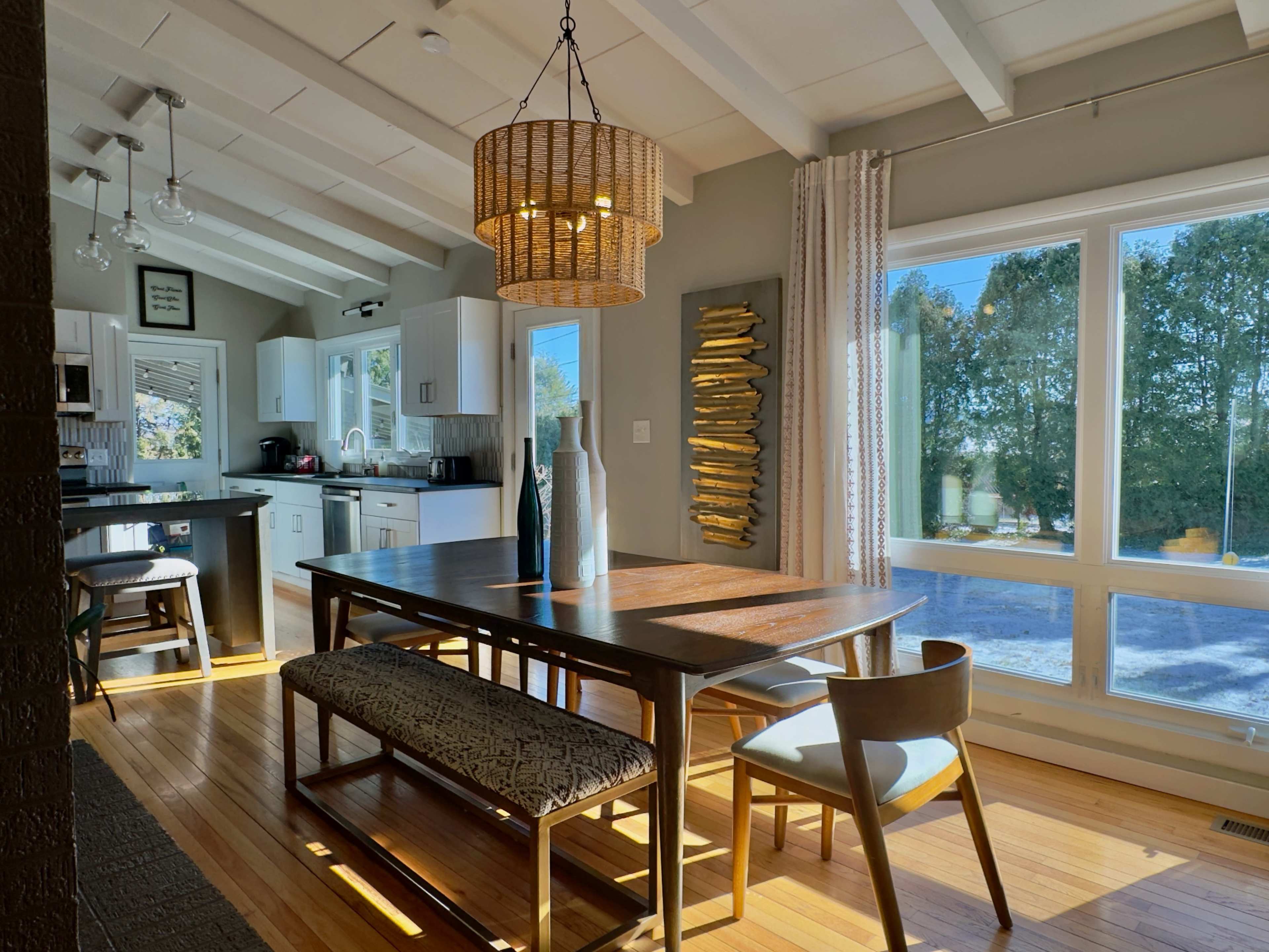 The image shows a modern dining area with a wooden table, a woven lamp fixture, and large windows allowing natural light to enter the space.