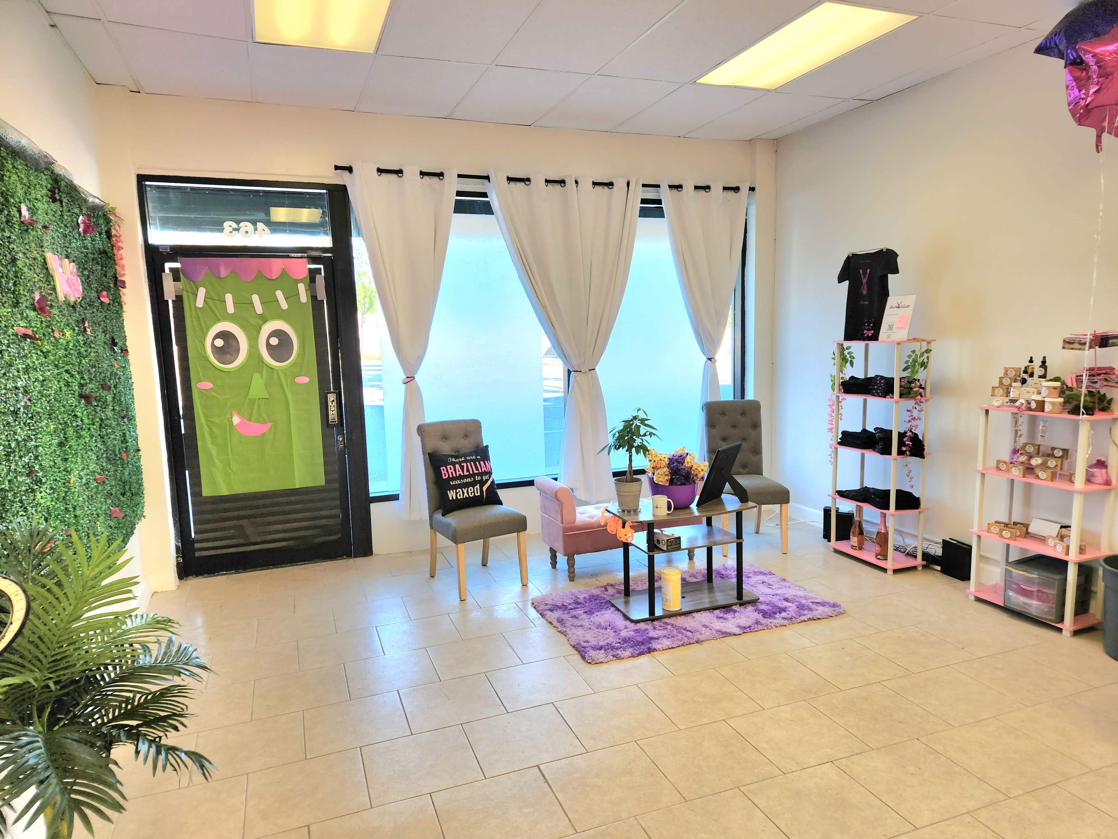 A brightly lit shop interior with two chairs, a table, a small plant, and shelves displaying various products, complemented by decorative elements like curtains and a wall covered in greenery.