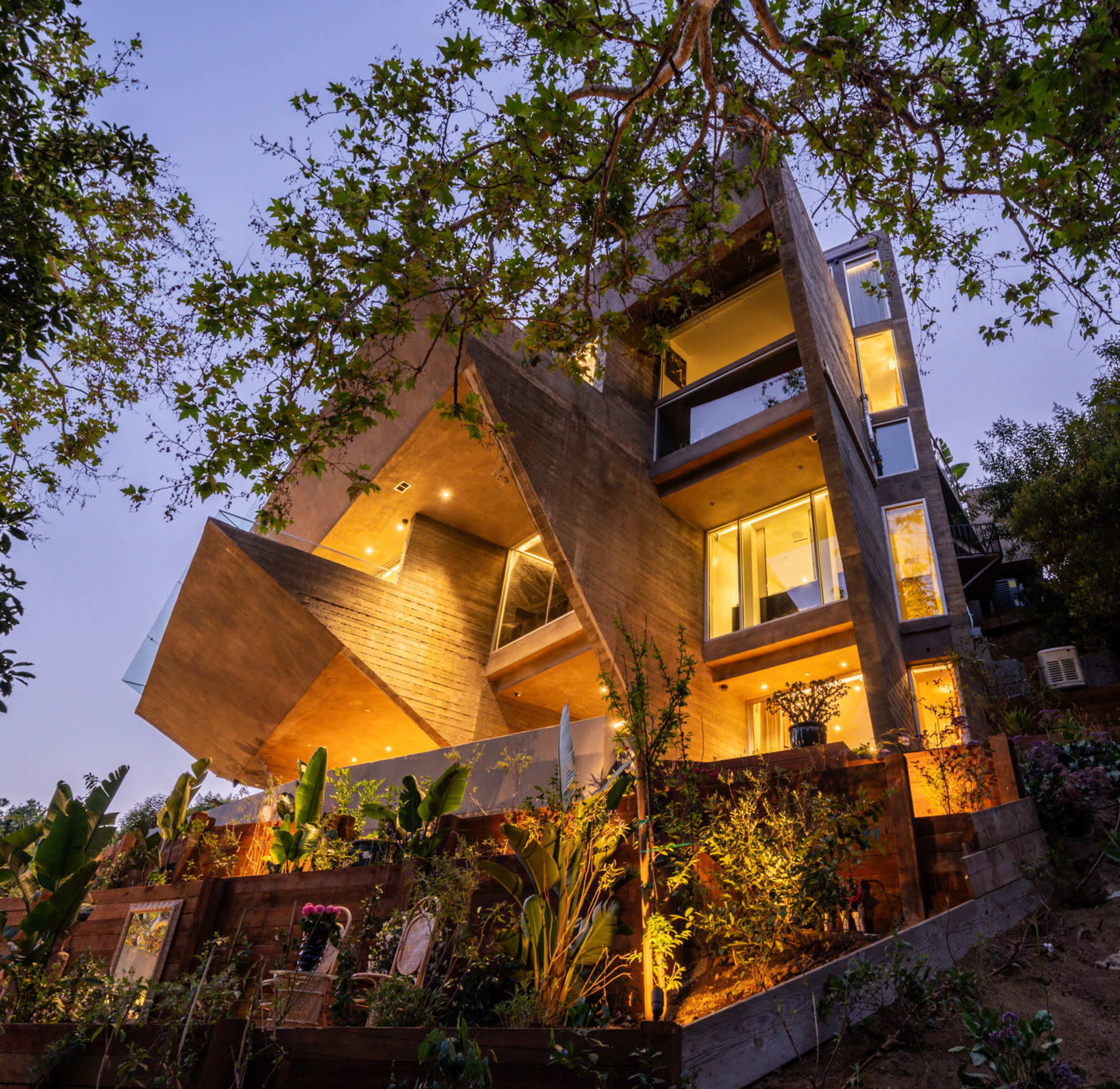 The image shows a modern angular concrete house surrounded by greenery, illuminated by warm outdoor lighting at dusk.