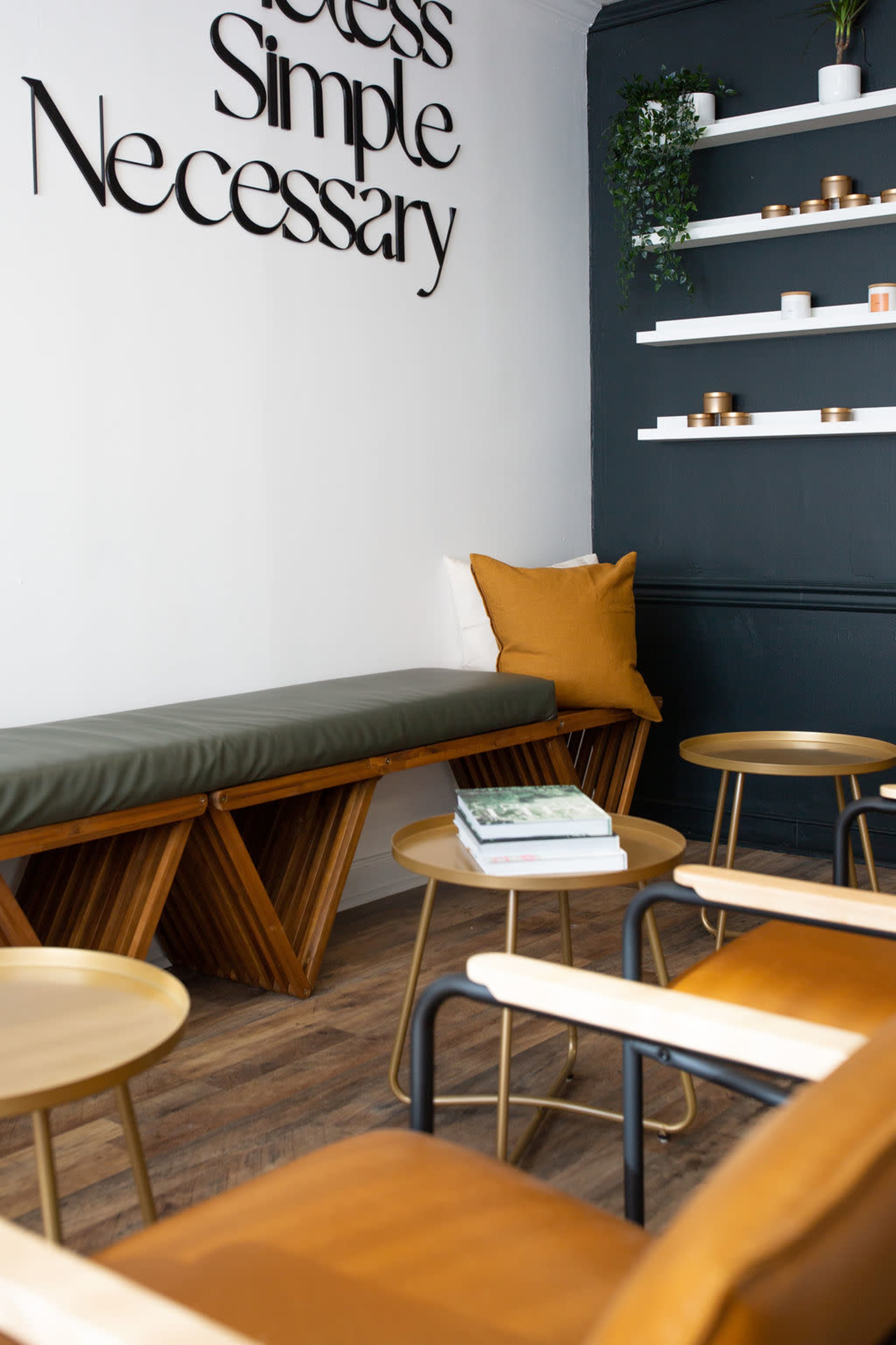 A minimalist seating area features a wooden bench with a green cushion, surrounded by small gold tables and a wall shelf displaying various items.