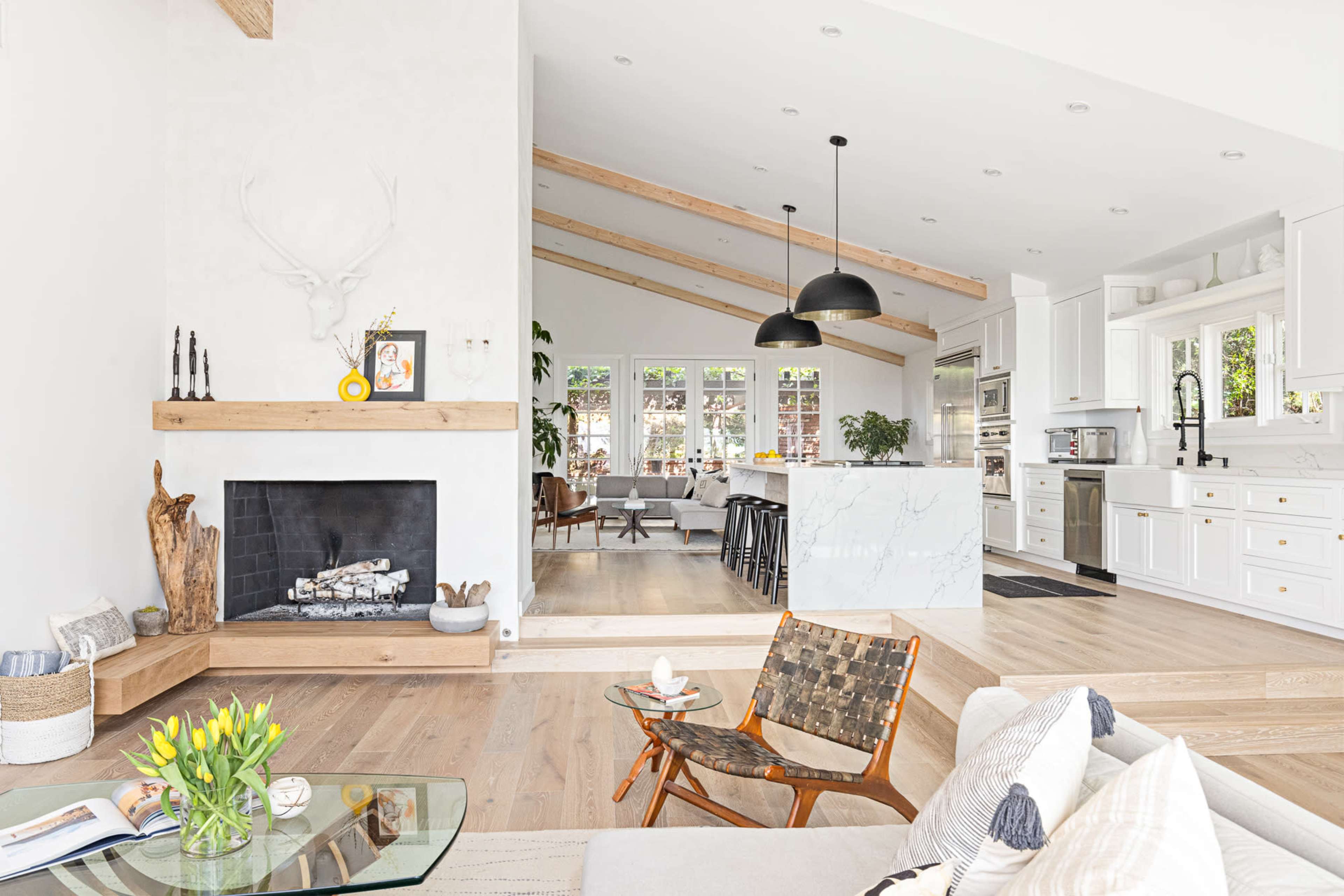 A spacious living area features a fireplace with a wooden mantel, a modern kitchen in the background, and large windows allowing natural light to fill the room.