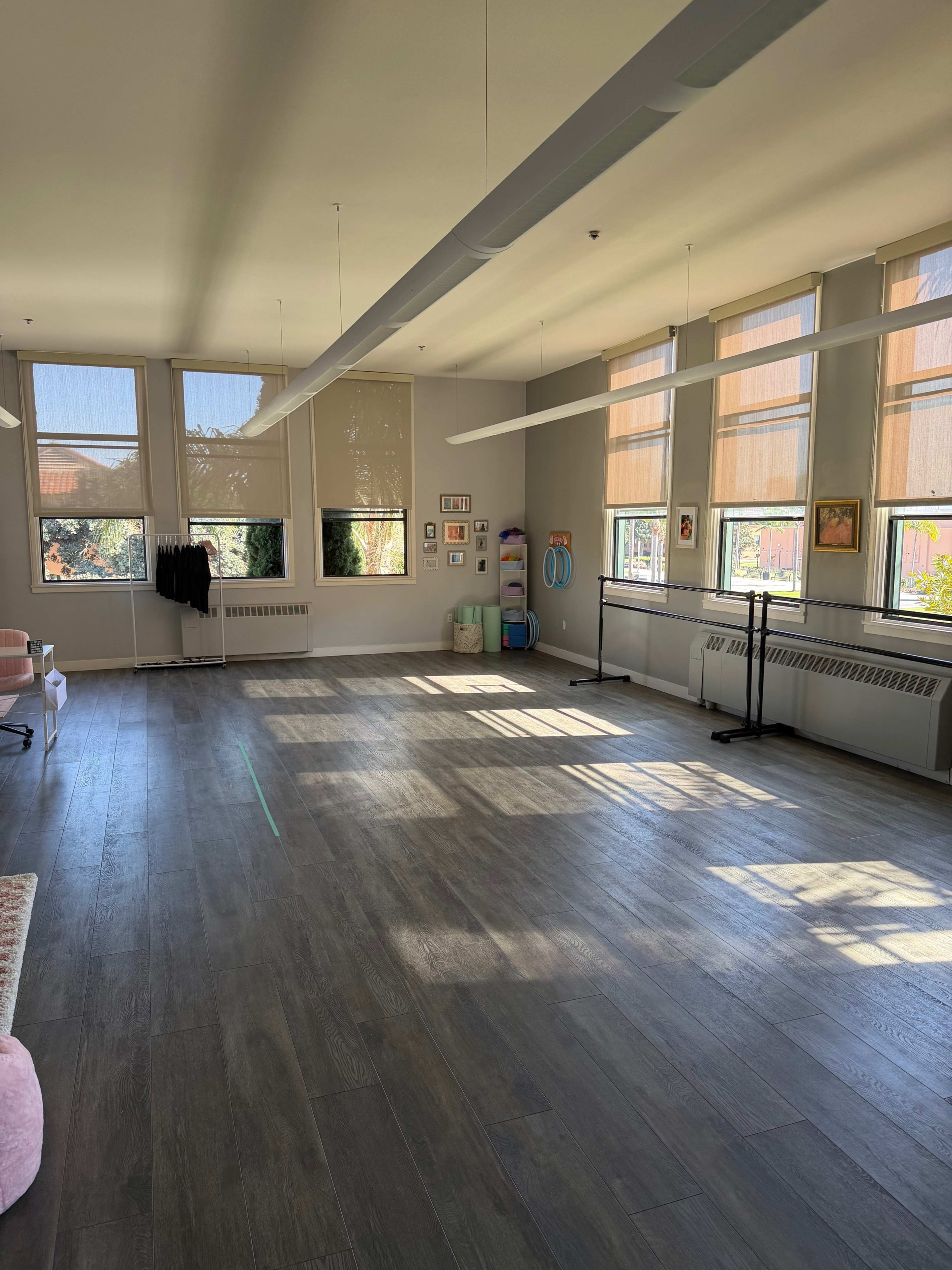 The image shows a spacious, empty room with wooden floors, large windows allowing natural light, and minimal furniture along the walls.