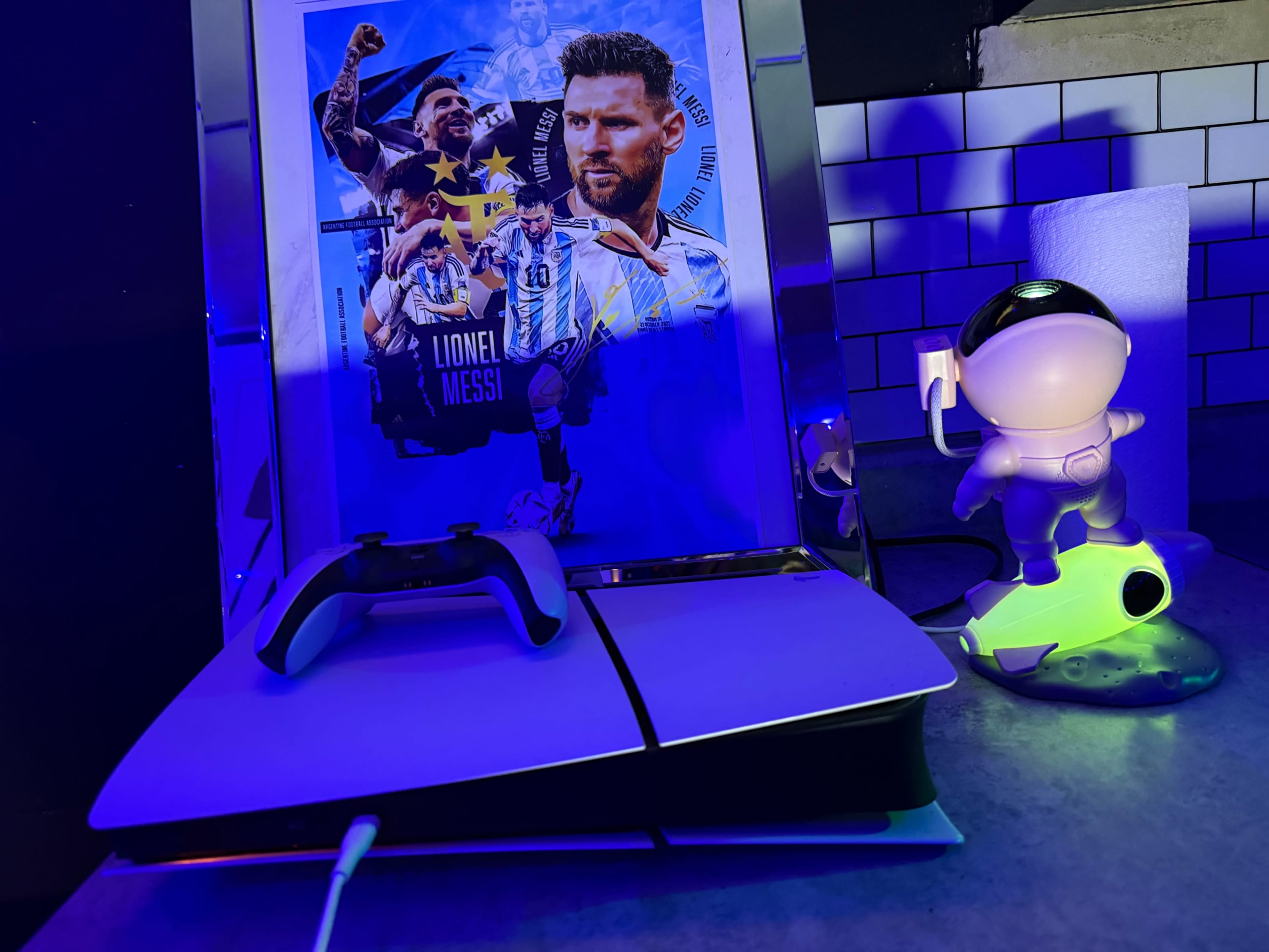 The image shows a PlayStation 5 console accompanied by a gaming controller, a framed poster of Lionel Messi, and a figurine illuminated with blue and green light.