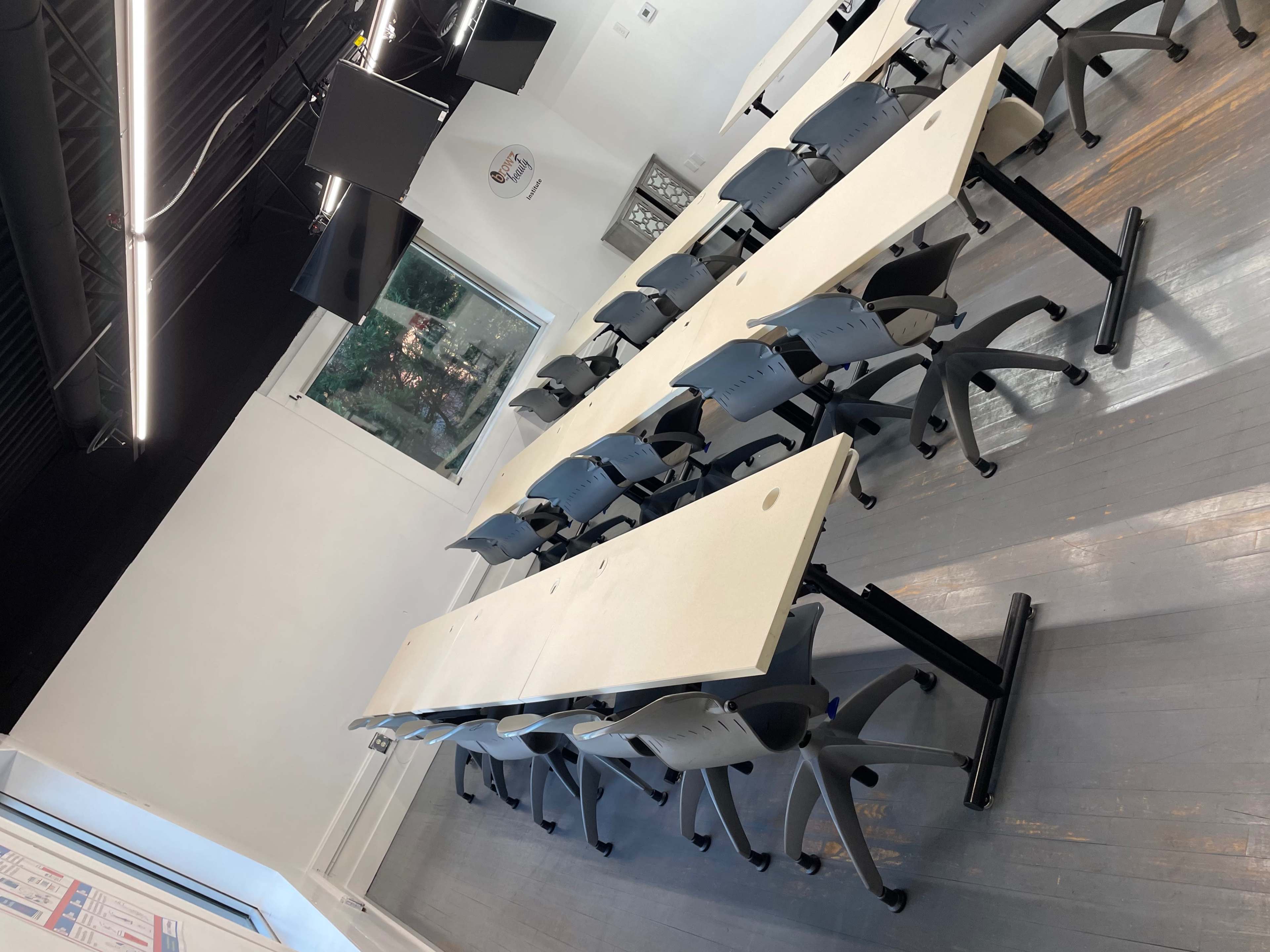 A classroom setting features long tables arranged in a U-shape with multiple chairs and several monitors mounted on the wall.