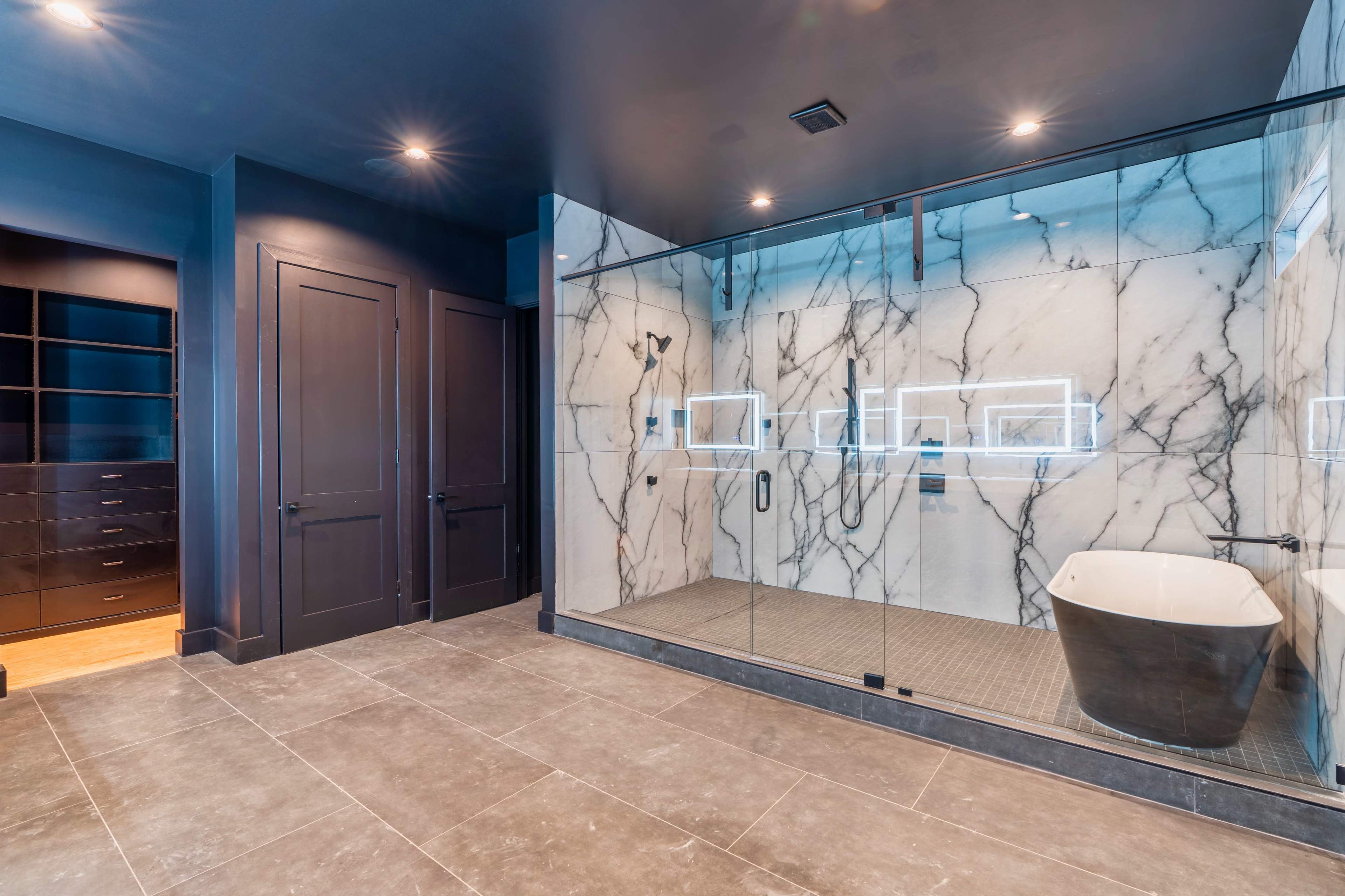 A modern bathroom featuring a glass-encased shower with marble walls, a freestanding black tub, and dark cabinetry.