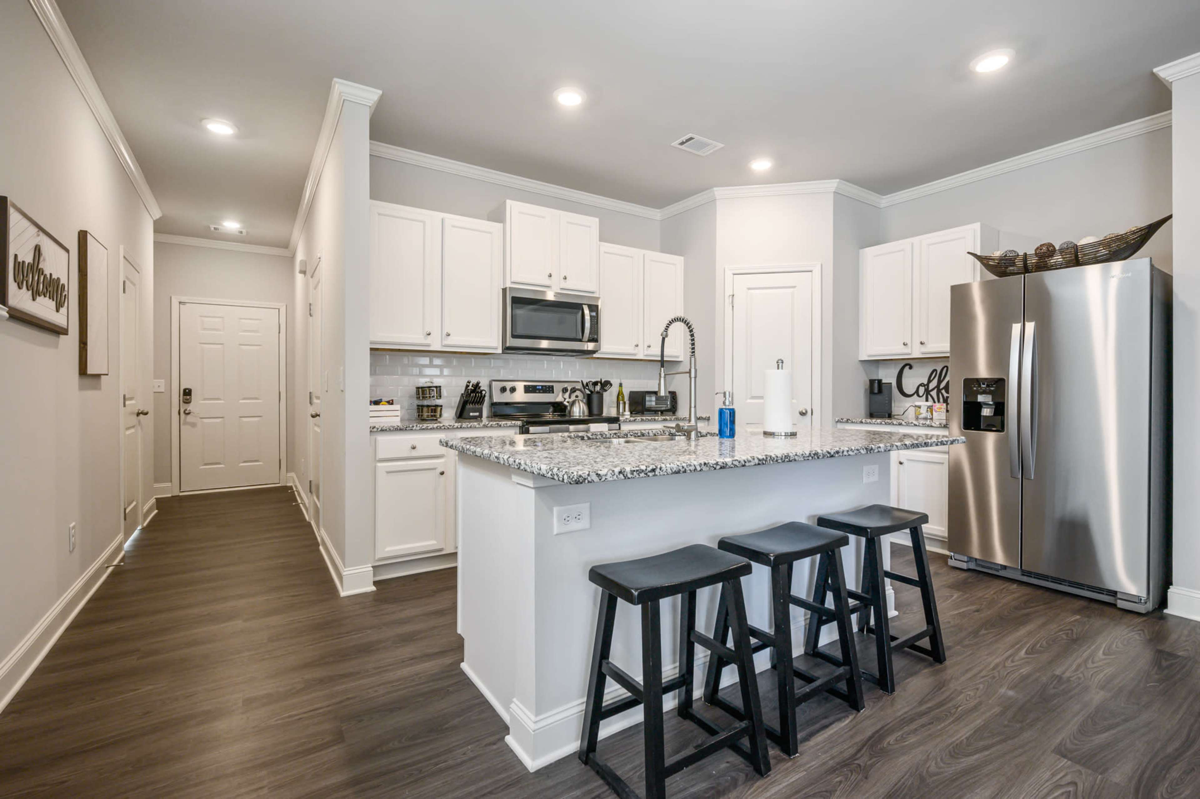 A modern kitchen with white cabinets, stainless steel appliances, and a granite countertop with four black stools at the island.