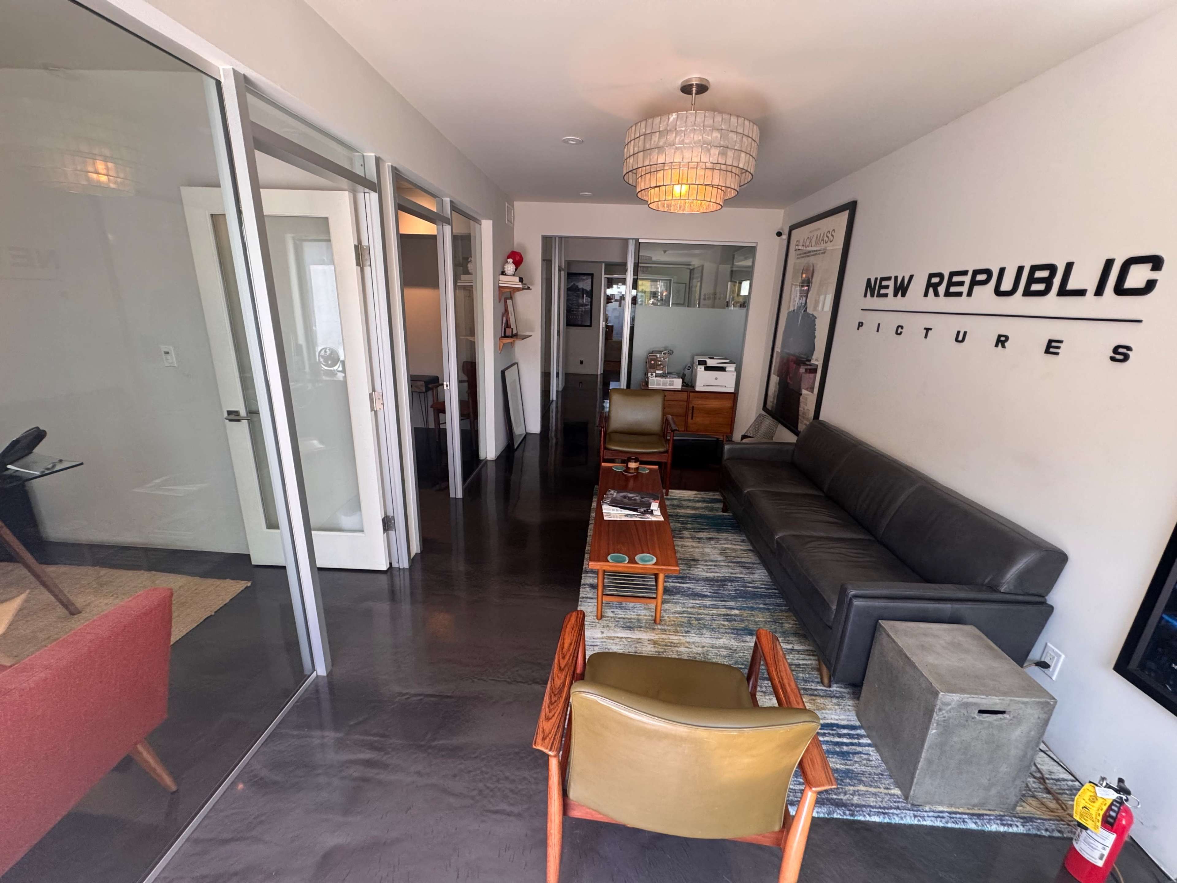 The image shows a modern office interior with a black sofa, wooden chairs, and a stylish chandelier, along with glass walls separating different rooms.
