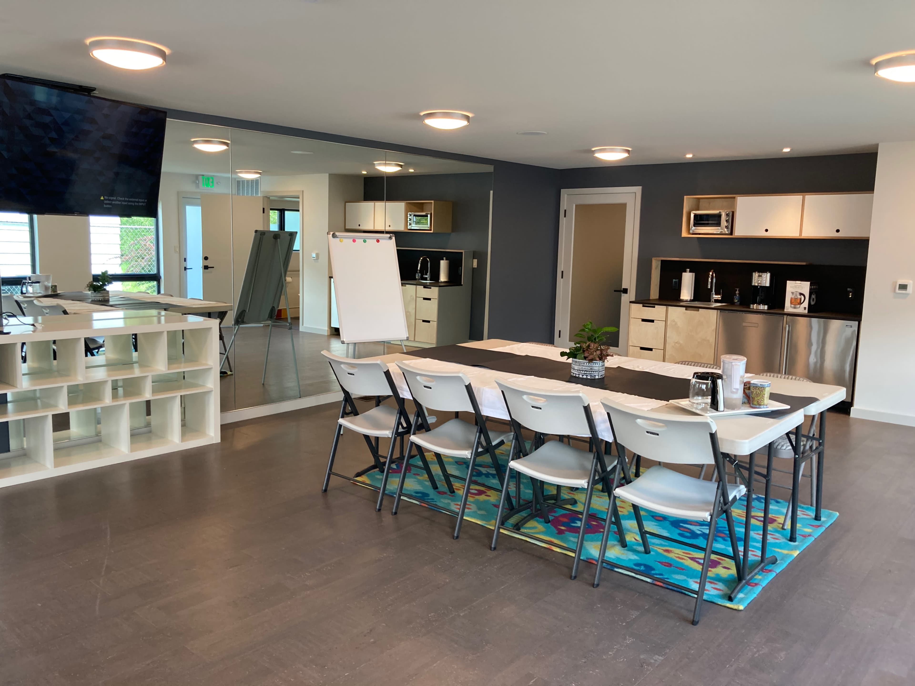 A modern meeting room with a large table surrounded by chairs, a kitchenette area, and a wall of mirrors reflecting the space.