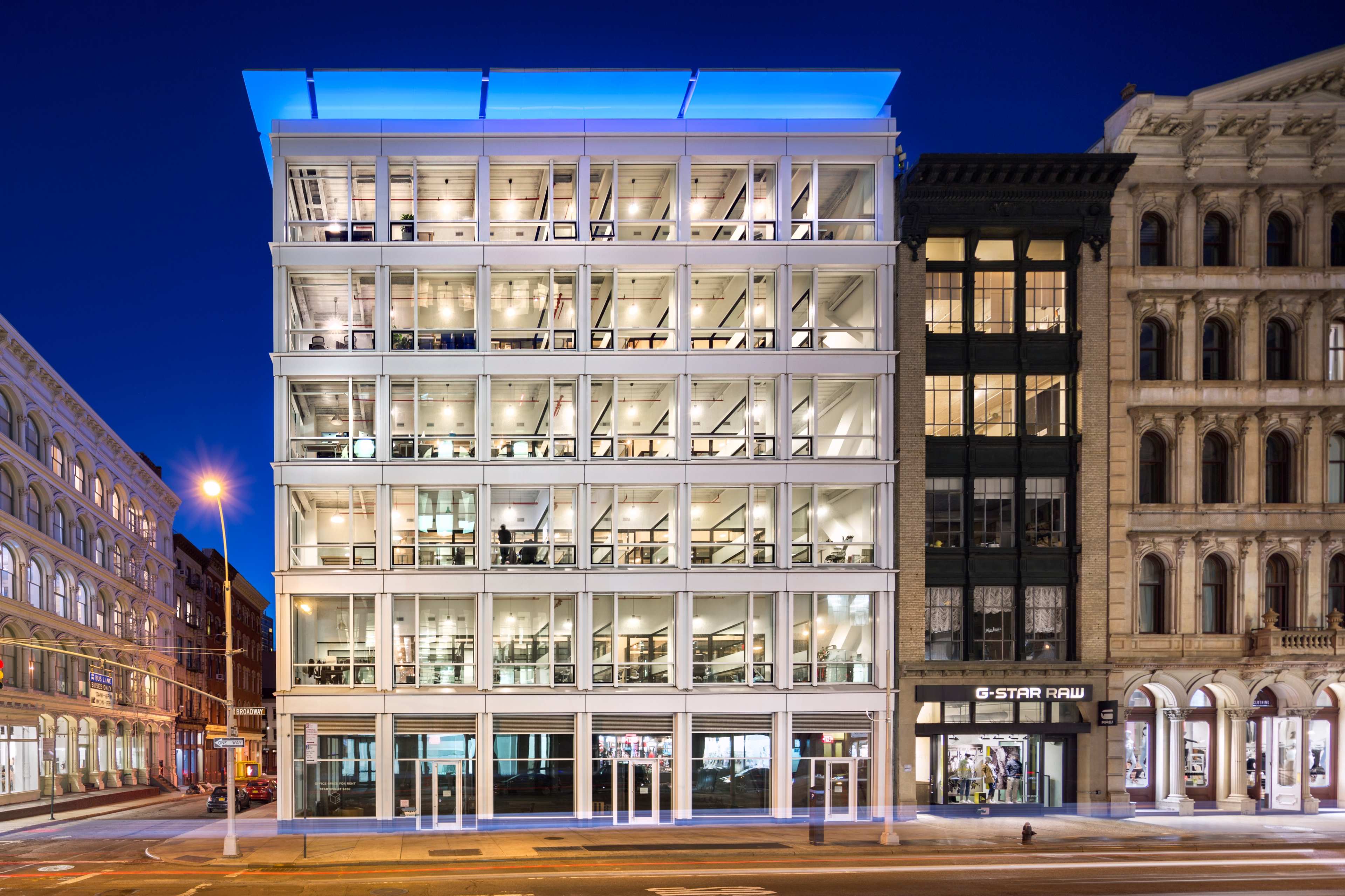 A modern, glass-fronted office building stands next to historic architecture in an urban setting at night.