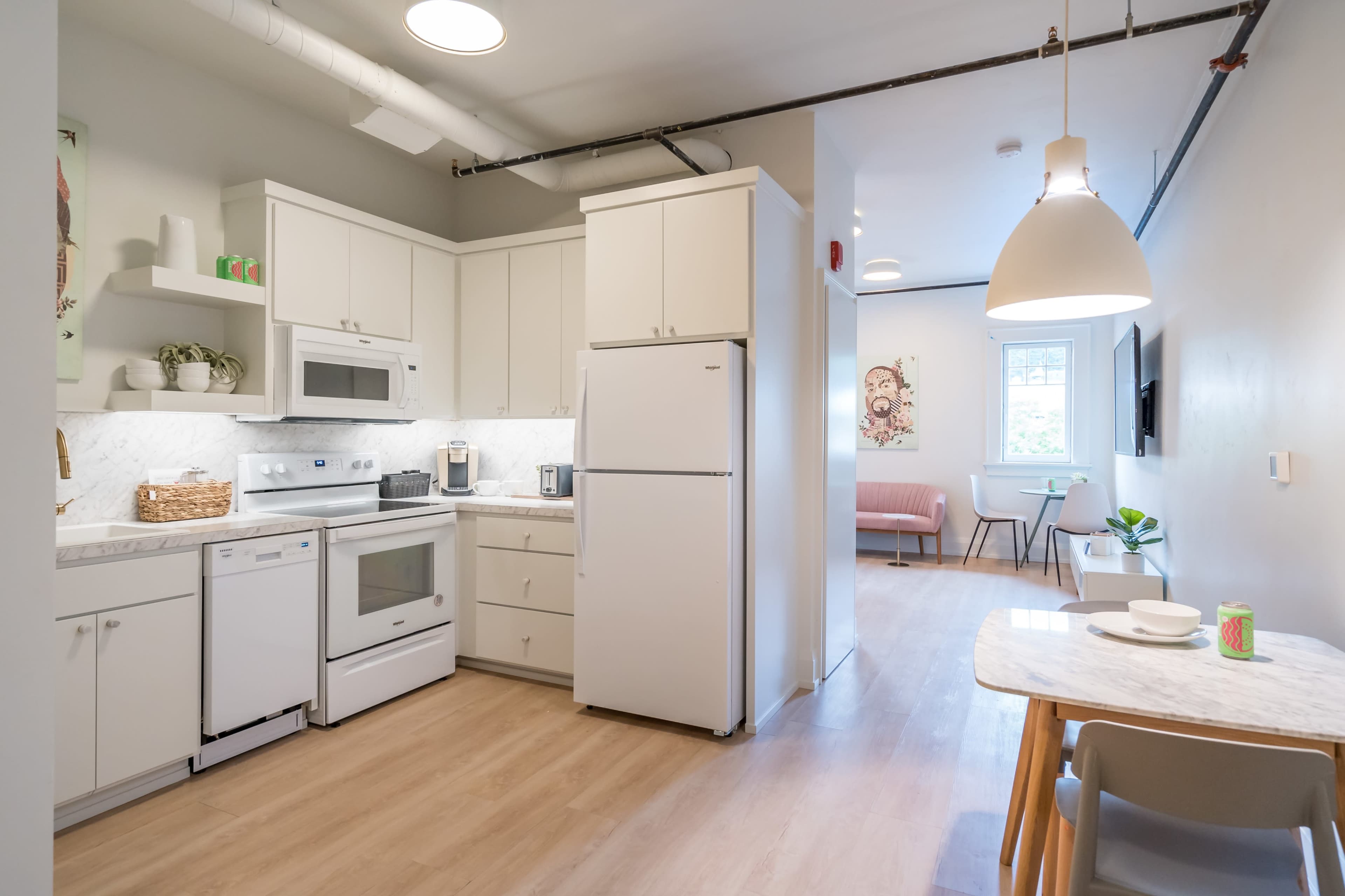 A modern kitchen with white cabinetry, a gray countertop, and stainless steel appliances, leading into a dining area with a pink chair and a small table.