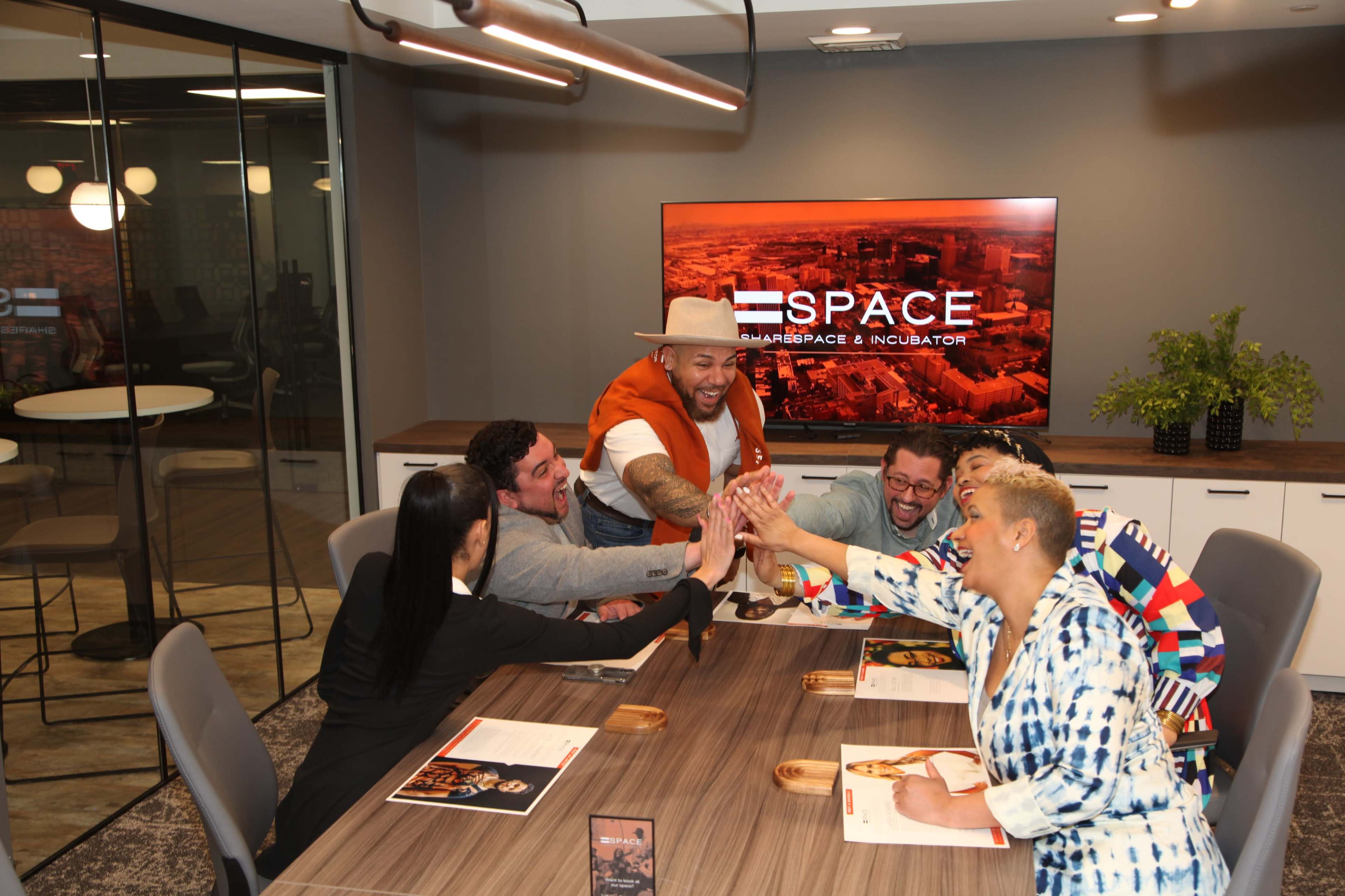 A diverse group of six people engage in a celebratory high-five around a conference table in a modern office setting.