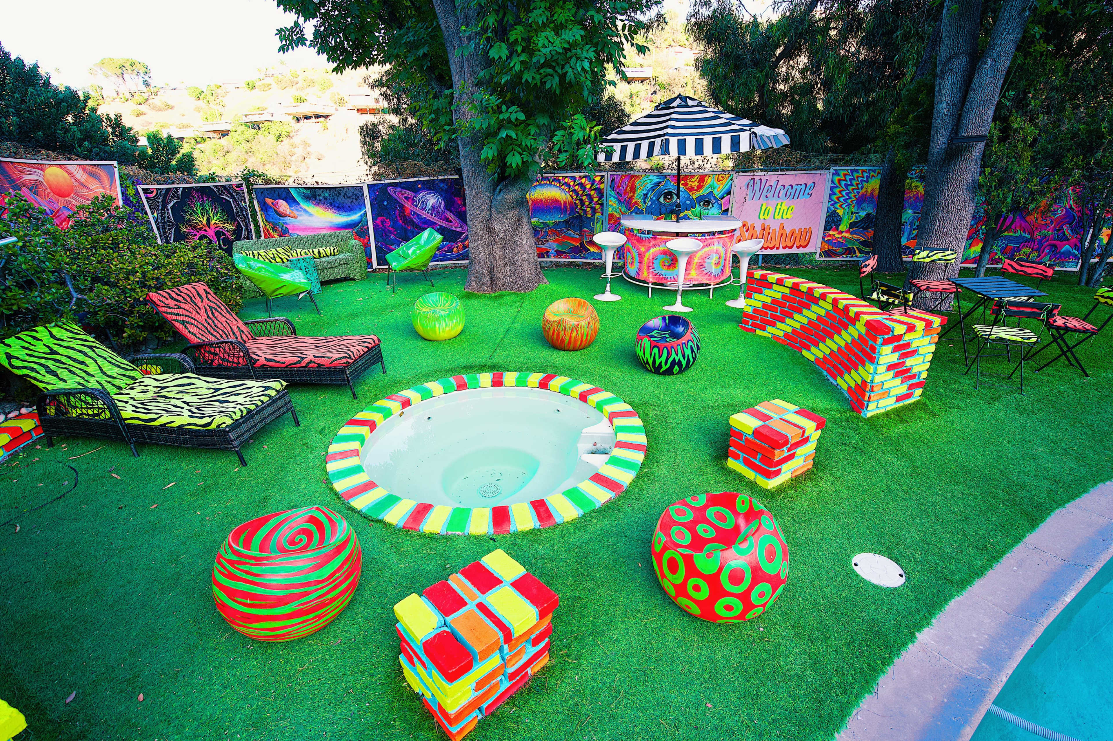 The scene features a vibrant outdoor area with colorful seating options, a small jacuzzi, and decorative spherical objects on green grass, surrounded by painted walls and a striped canopy.