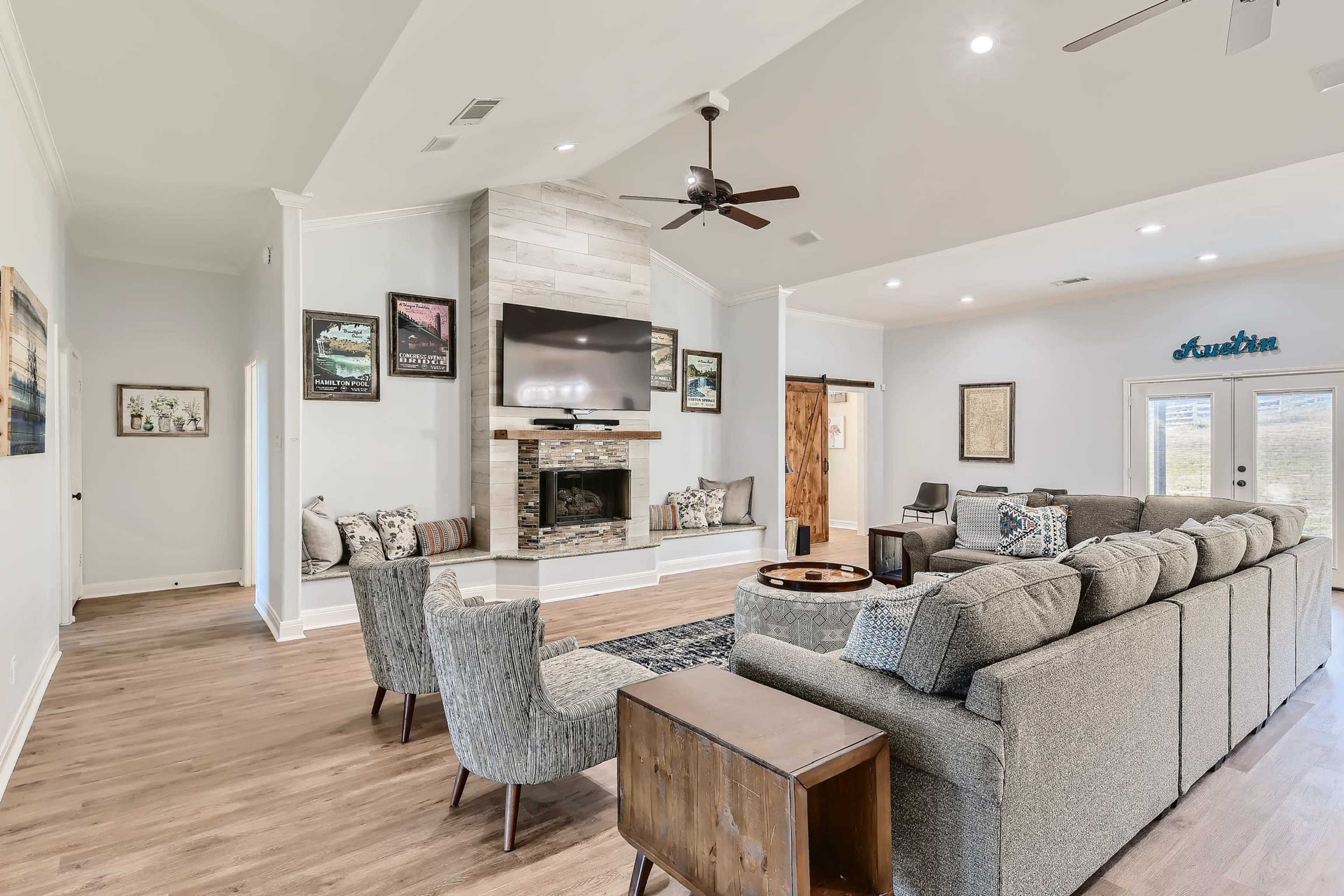 The image shows a modern living room featuring a fireplace with a mounted television, comfortable seating, and large windows allowing natural light.