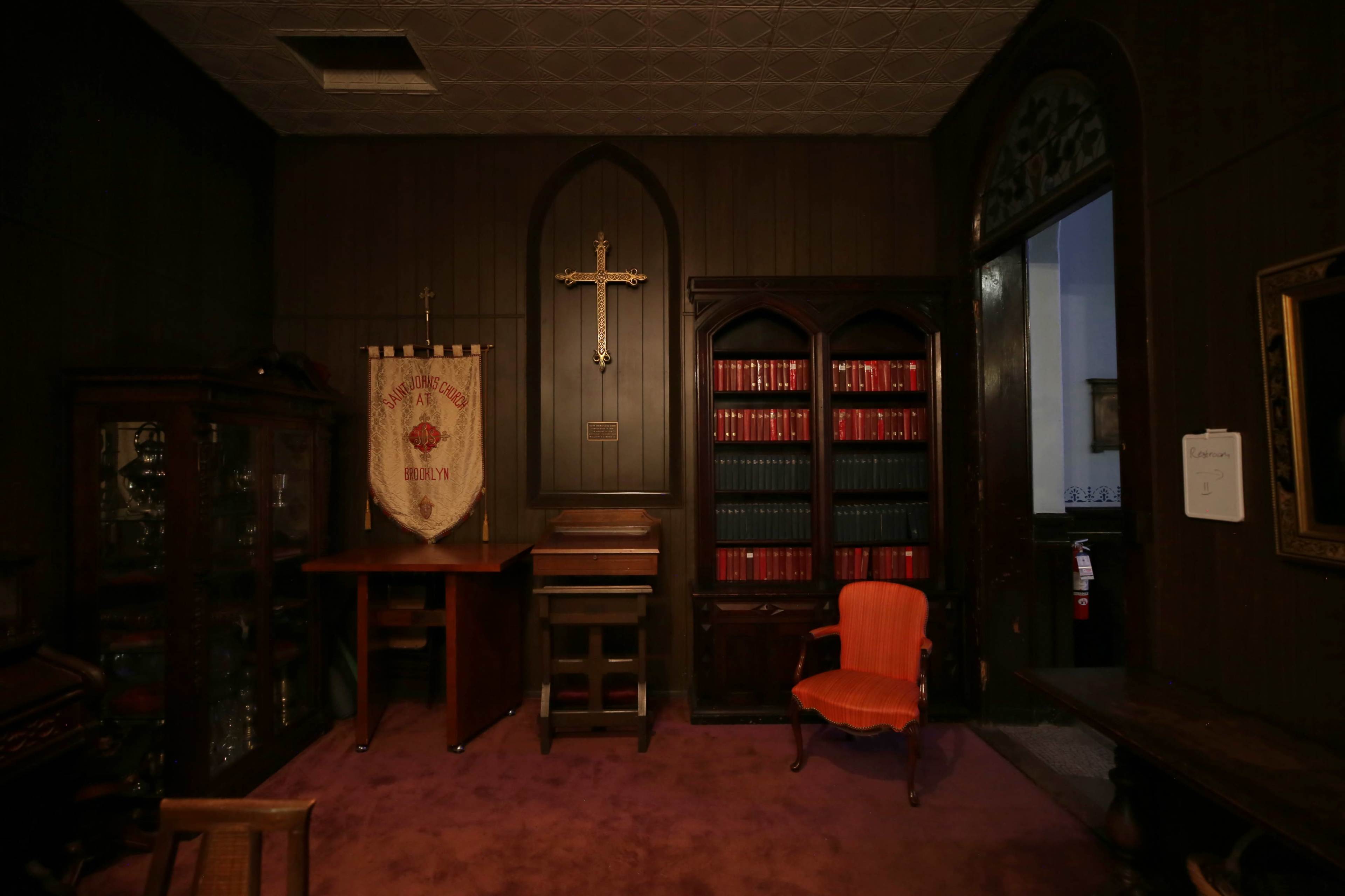 The room features a wooden decor with a cross on the wall, a bookshelf filled with red volumes, a decorative banner, and an orange chair.